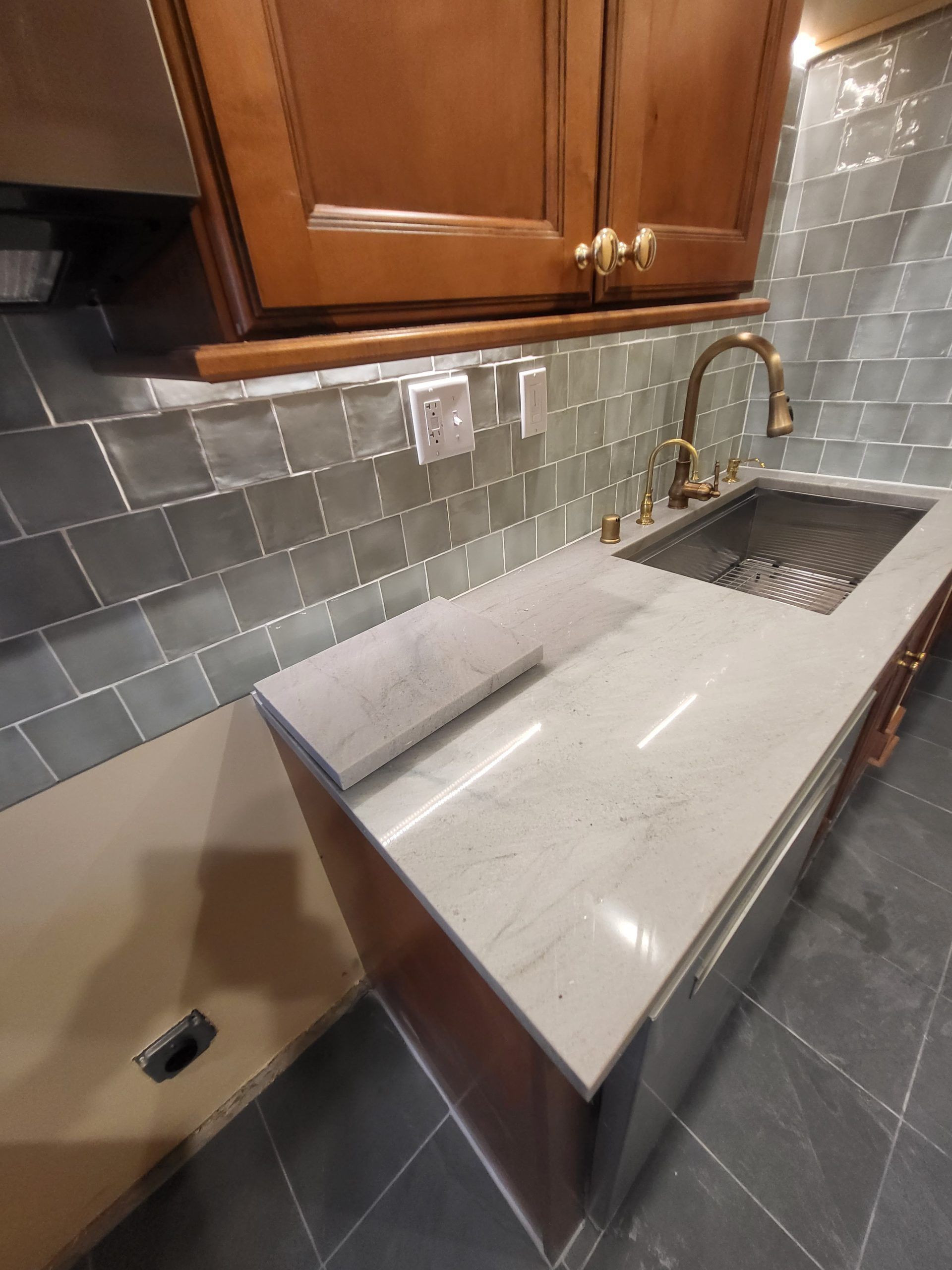 Kitchen with light countertops, dark backsplash, wood cabinets, and brass faucet.