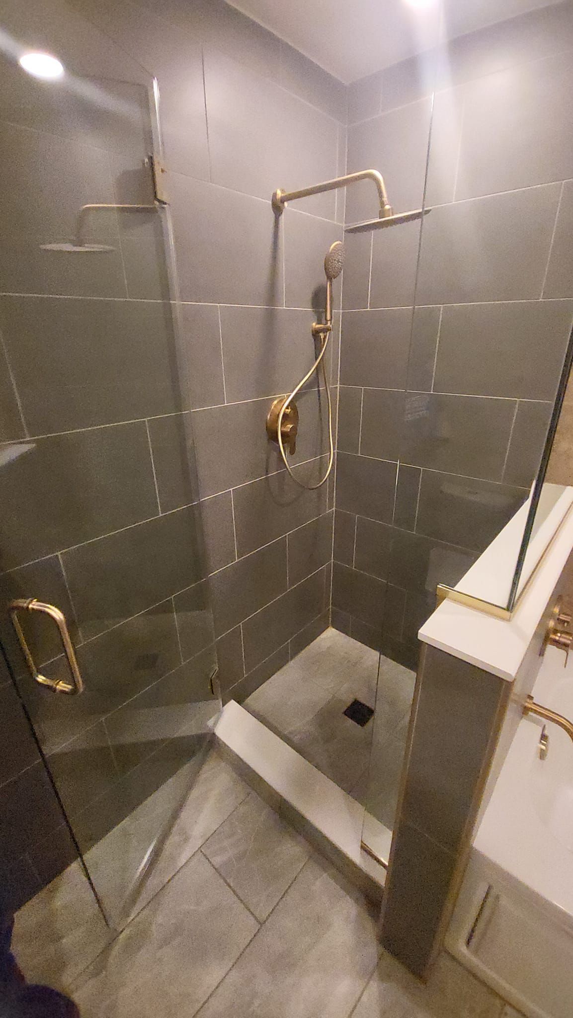 Gray-tiled shower with gold fixtures, glass door, and a white, angular ledge.