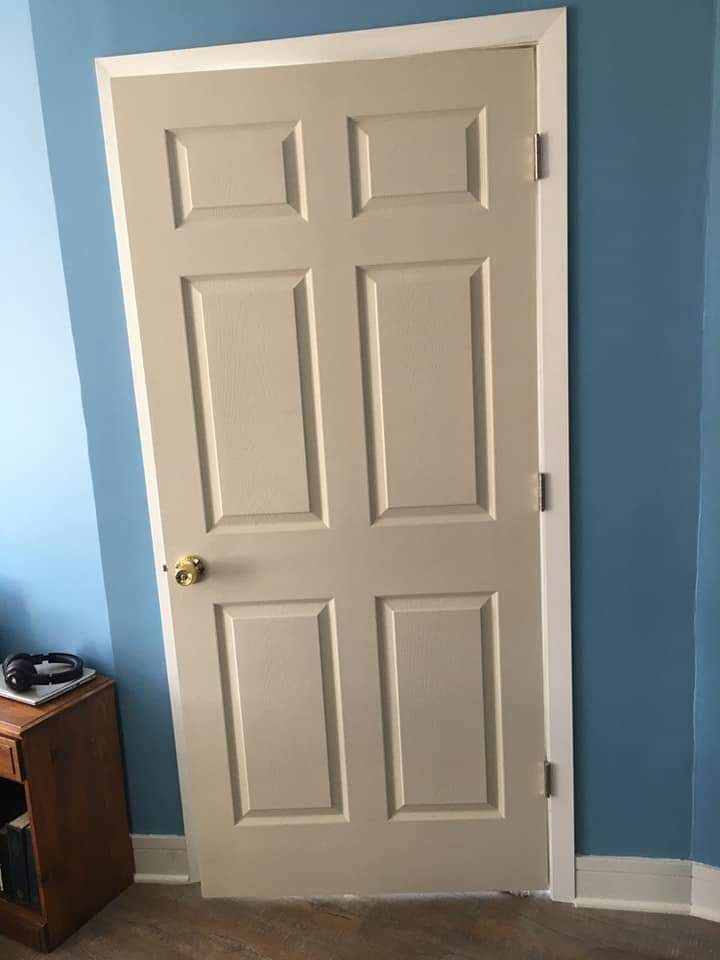 Beige six-panel door slightly ajar within white trim, leaning against blue walls and a wooden bedside table.