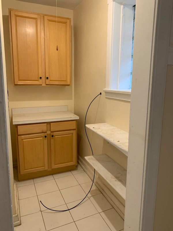 A utility room with cabinets, countertop, window, and shelving. Beige walls, and white tiled floor.