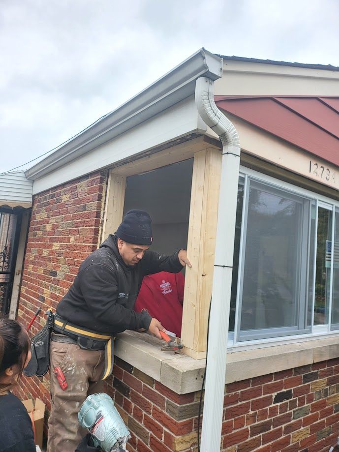 Two construction workers in a room with drywall, one applying material to the wall and the other inspecting a window.