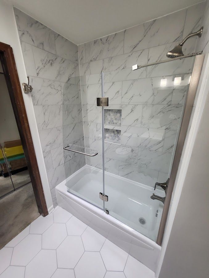 Bathroom with white tub, marble-look tiled walls, glass shower door, and hexagonal floor tiles.