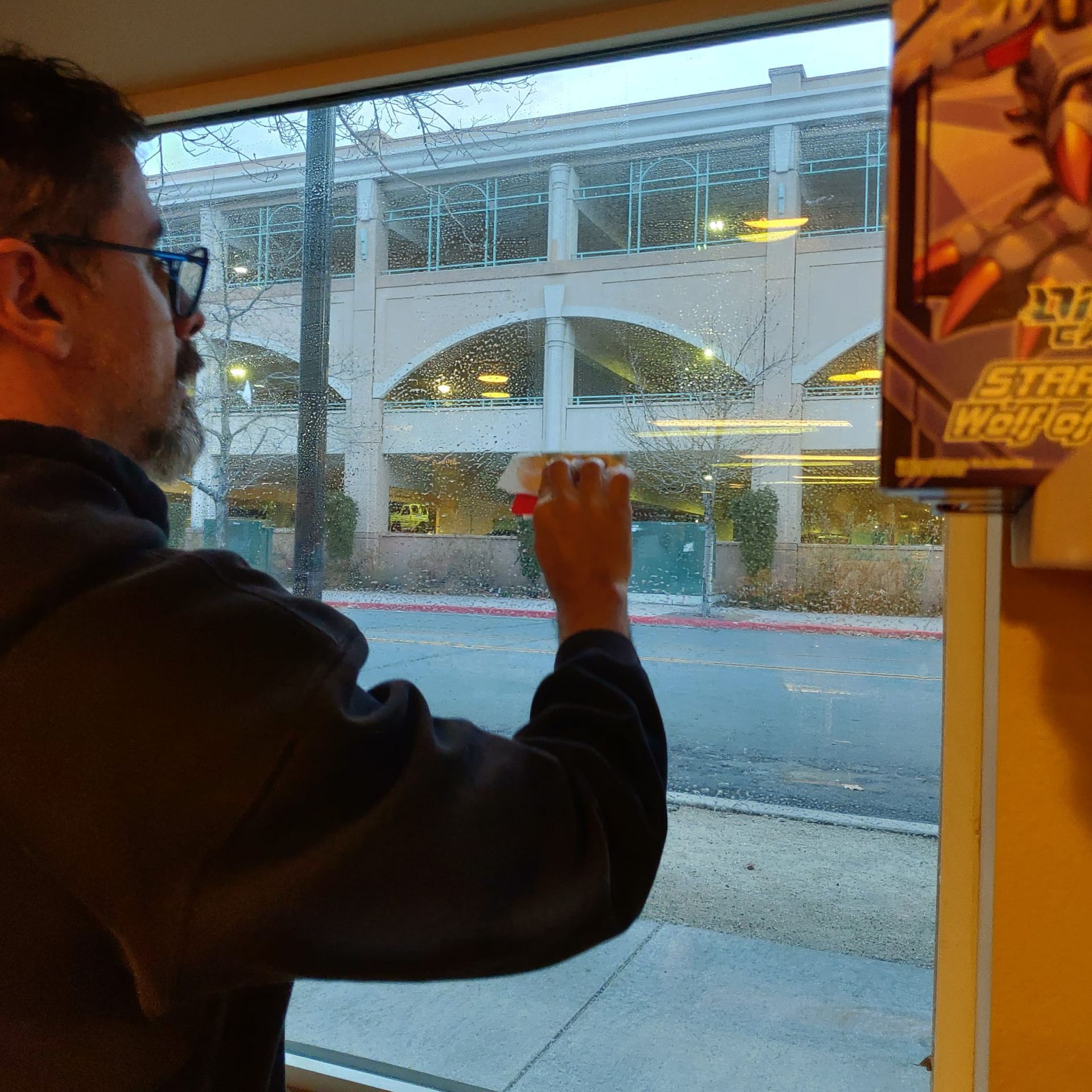 A person with a beard and glasses cleans a large window looking out onto a street and parking garage.