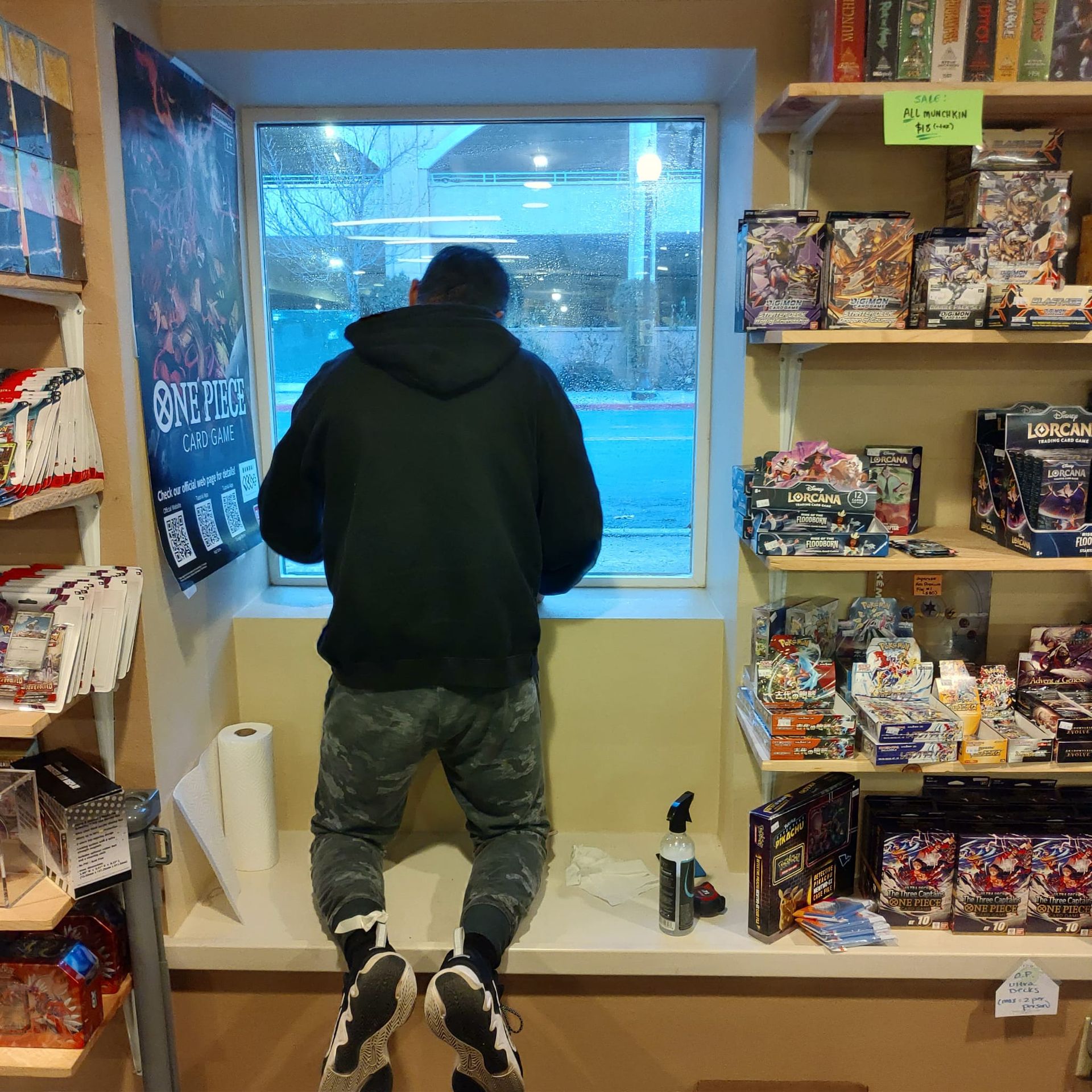 A person in a black hoodie and camo pants kneels on a ledge in a store, looking out a window between shelves of products.