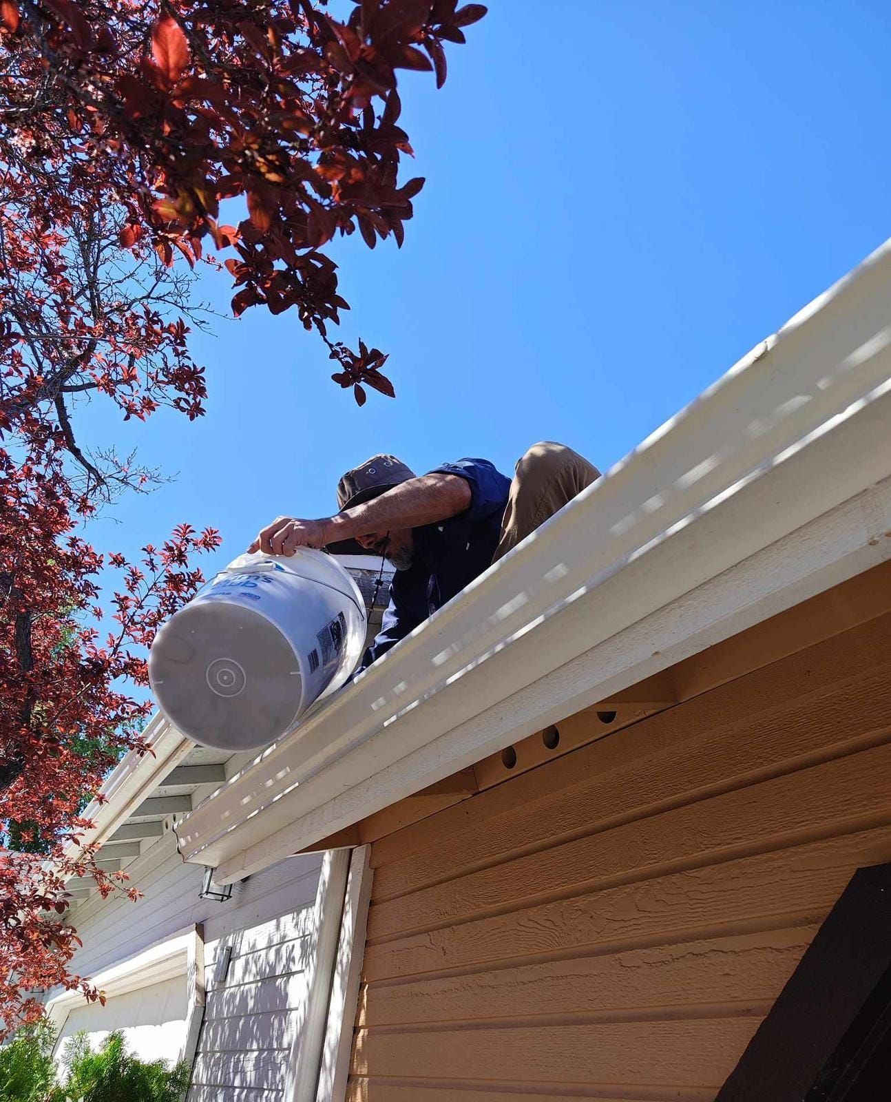 A person on a roof leans over to dump debris from a plastic bucket into the rain gutter.