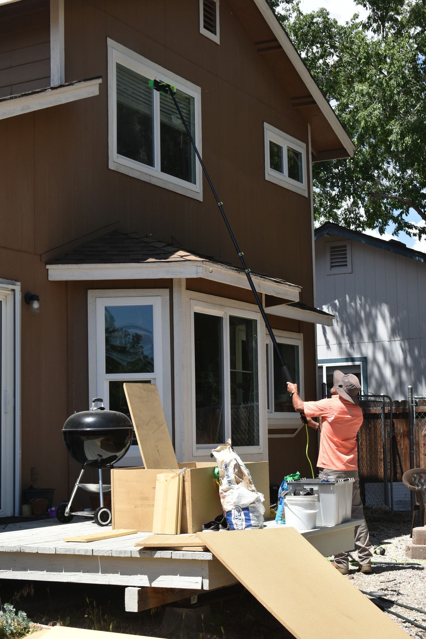 A person in an orange shirt uses a long pole to clean the second-story window of a brown house.