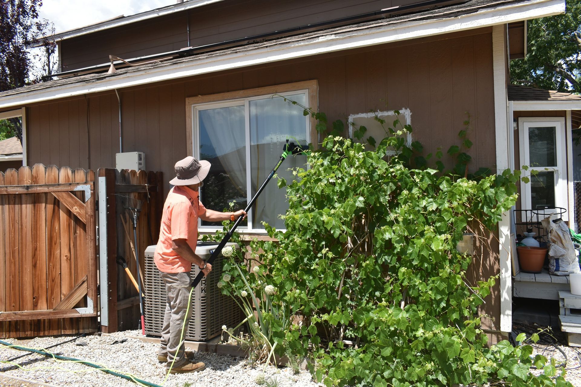 A person in a hat and orange shirt uses a long pole tool to clean a window on the side of a brown house.