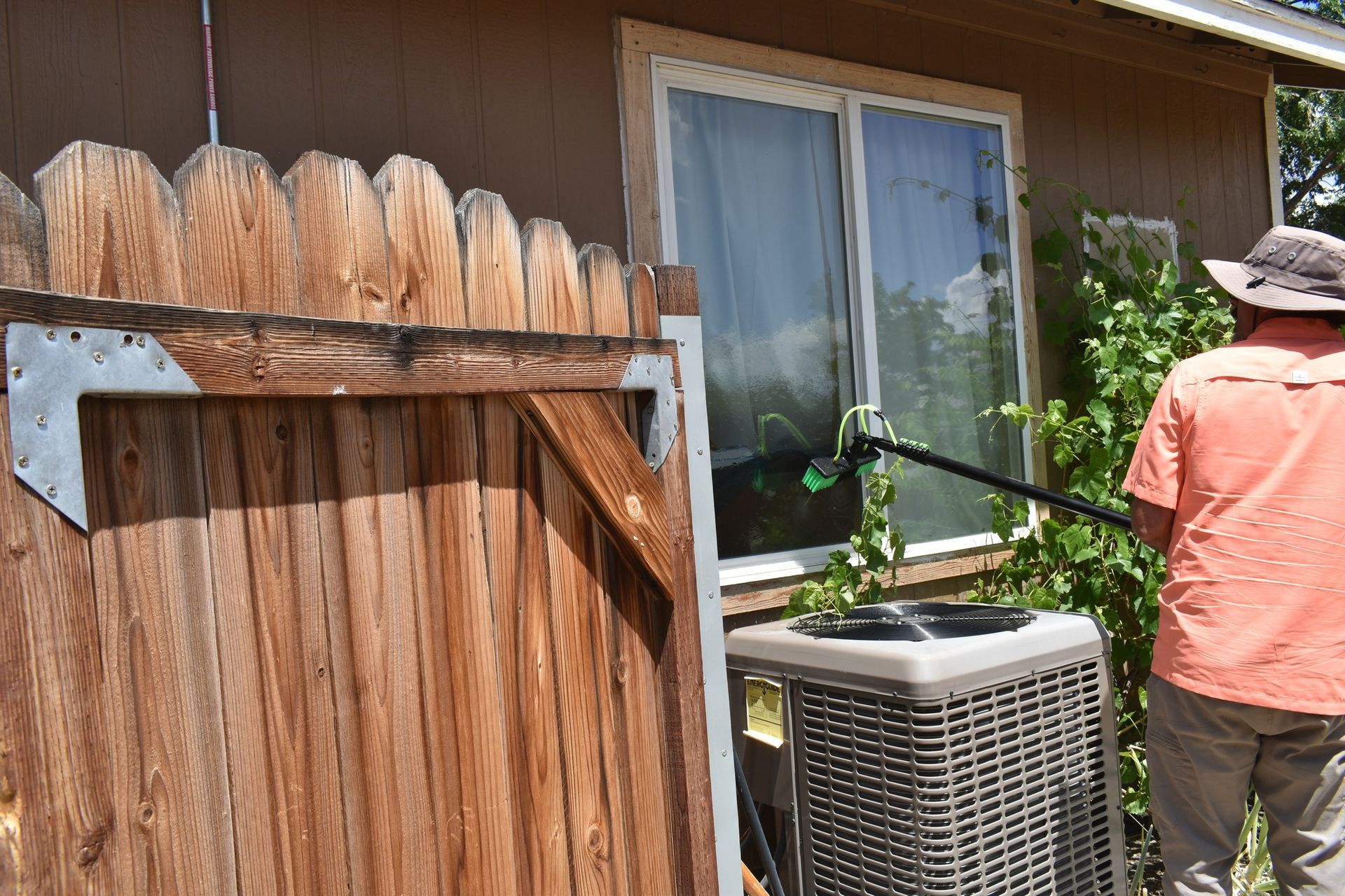 A person in an orange shirt uses a long-handled tool to clean the exterior of a window near a fence and AC unit.