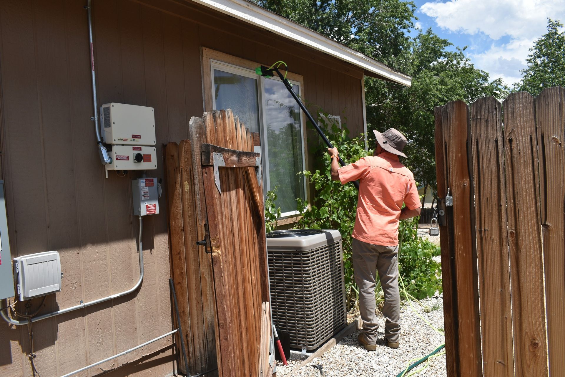 A person wearing a hat and orange shirt uses a long-handled tool to clean the exterior of a home near an air conditioner.