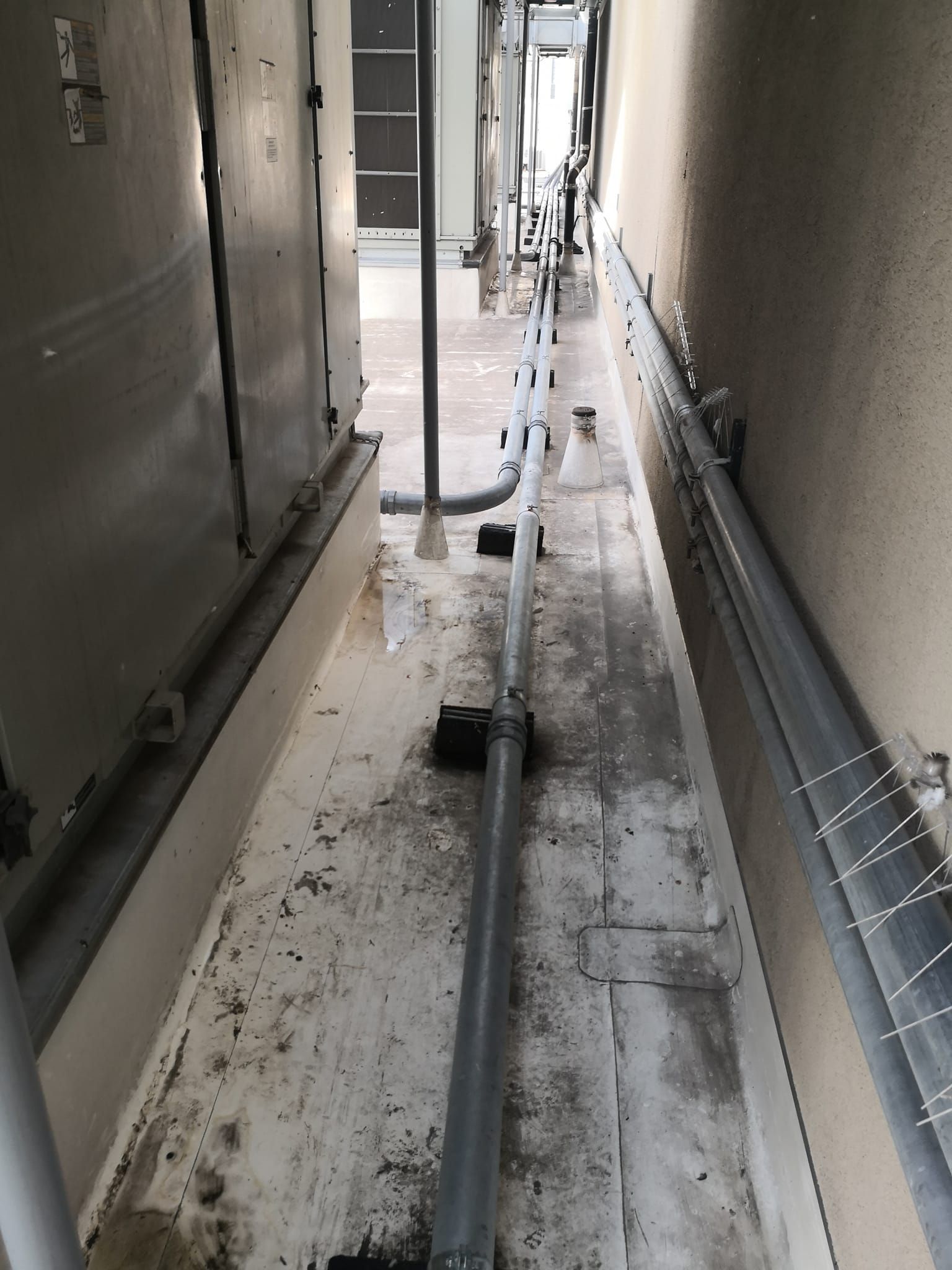 A narrow outdoor corridor with pipes running along the ground and wall between a metal structure and a concrete wall.