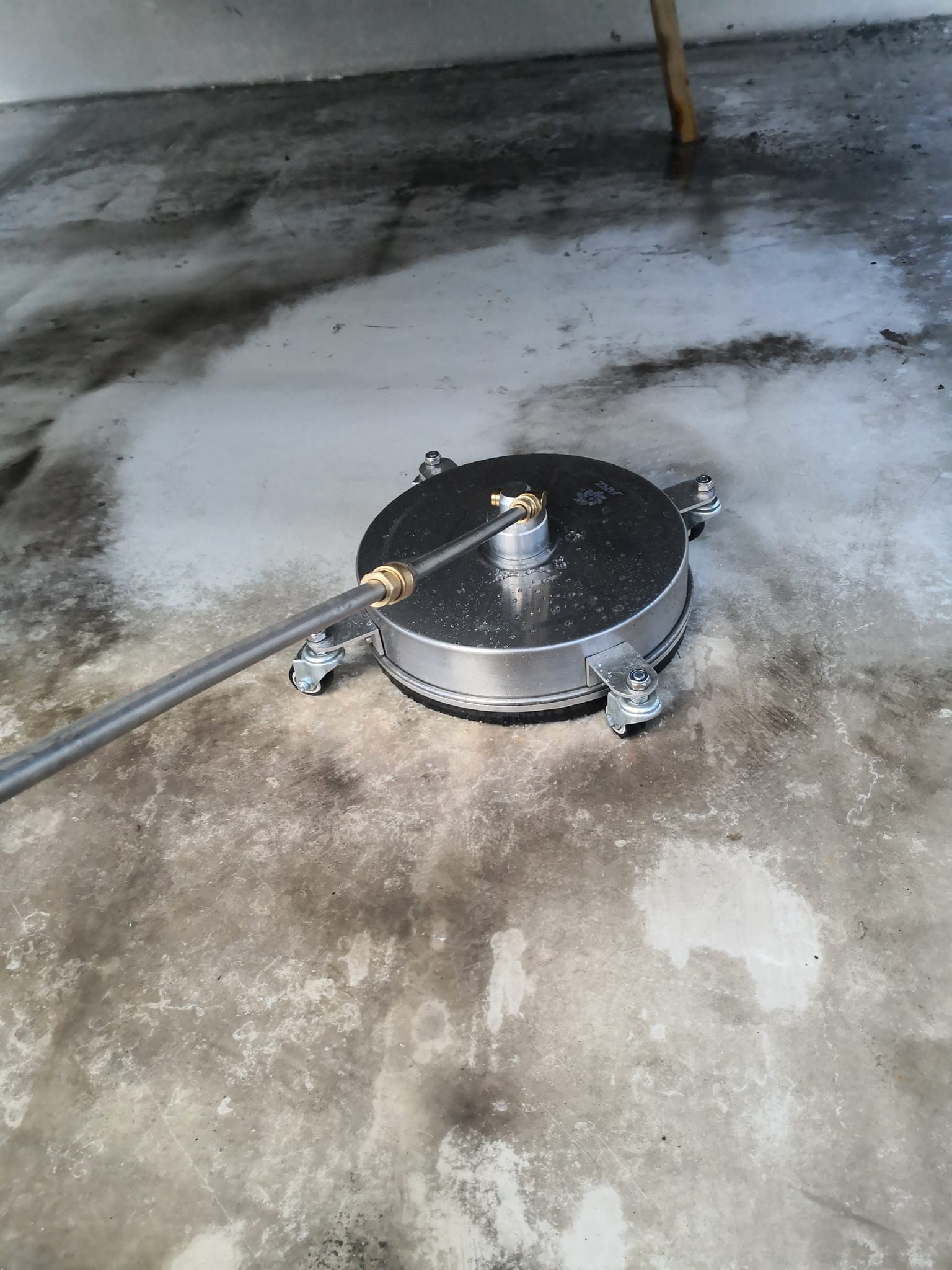 A circular metal pressure washer surface cleaner being used to clean a dirty concrete floor.