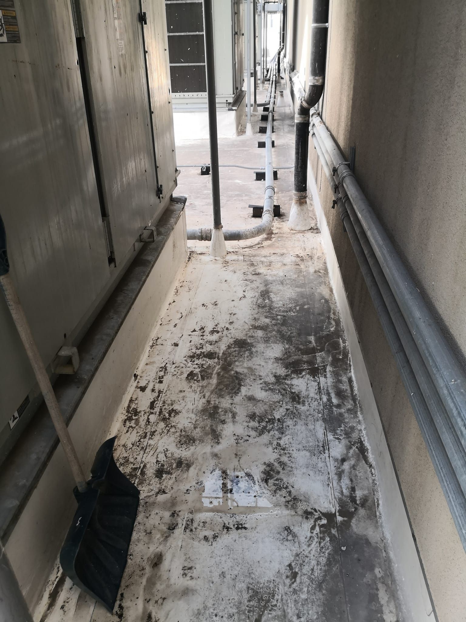 A narrow outdoor corridor between an industrial unit and a wall, with a shovel leaning against the soiled white floor.