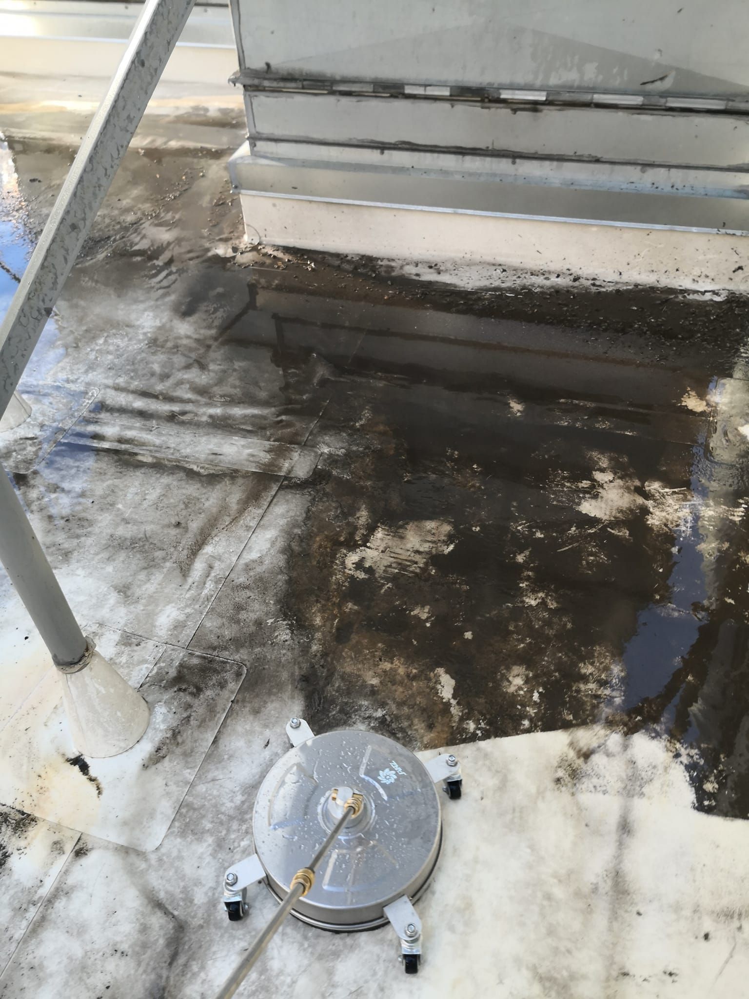 A circular pressure washer tool sits on a stained, flat roof surface next to a metal structure.