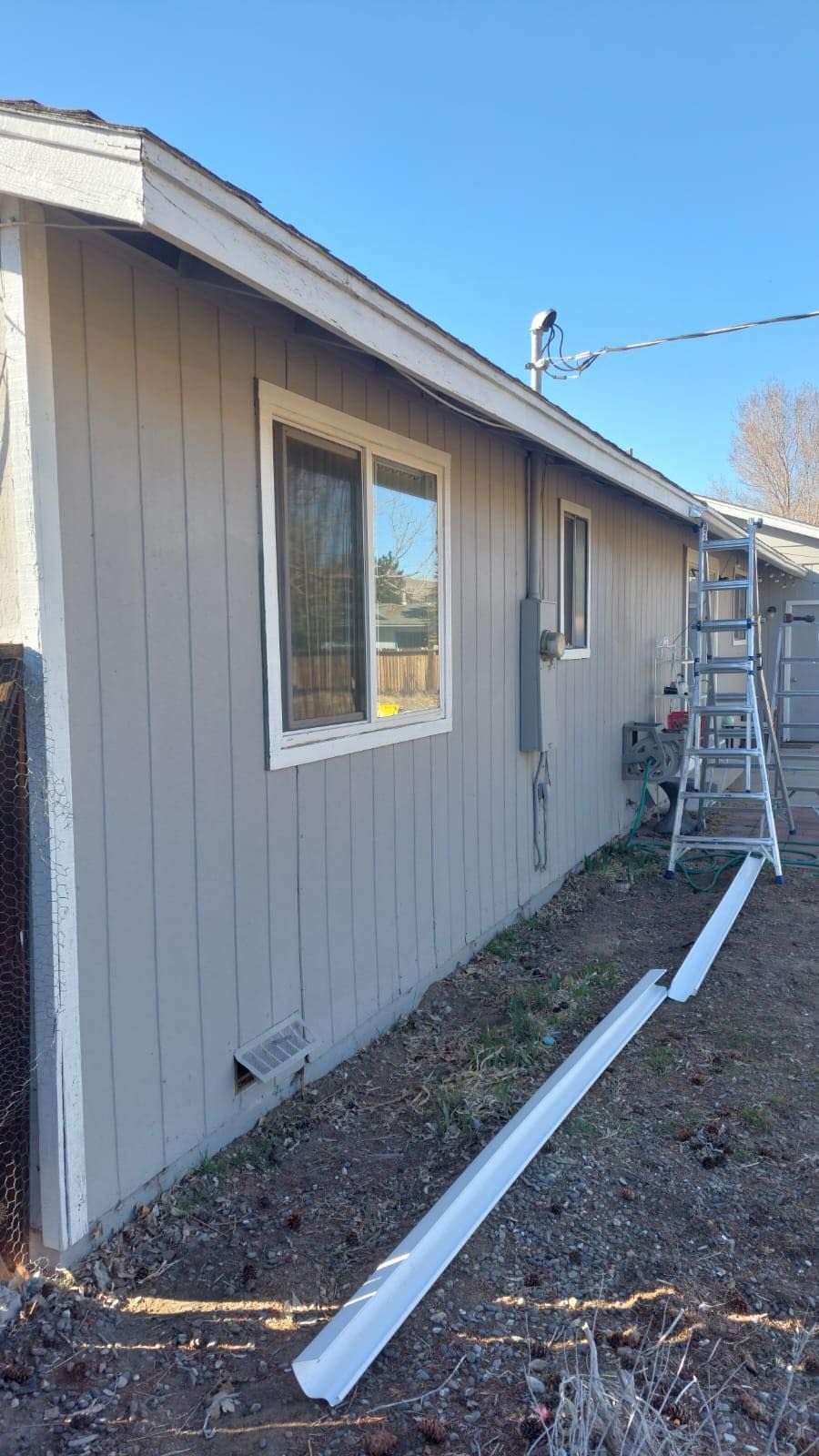 A tan house exterior with a window, an electrical box, and a ladder leaning against a wall with detached white gutters.