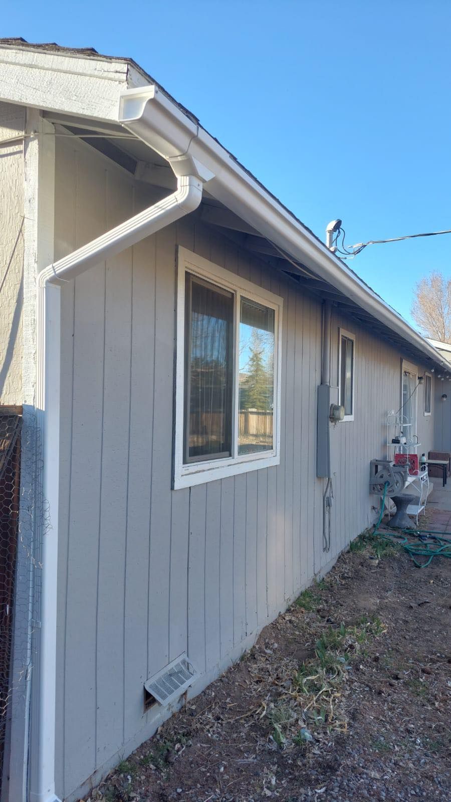 A side view of a light gray house featuring vertical siding, white window trim, and a white gutter under a blue sky.