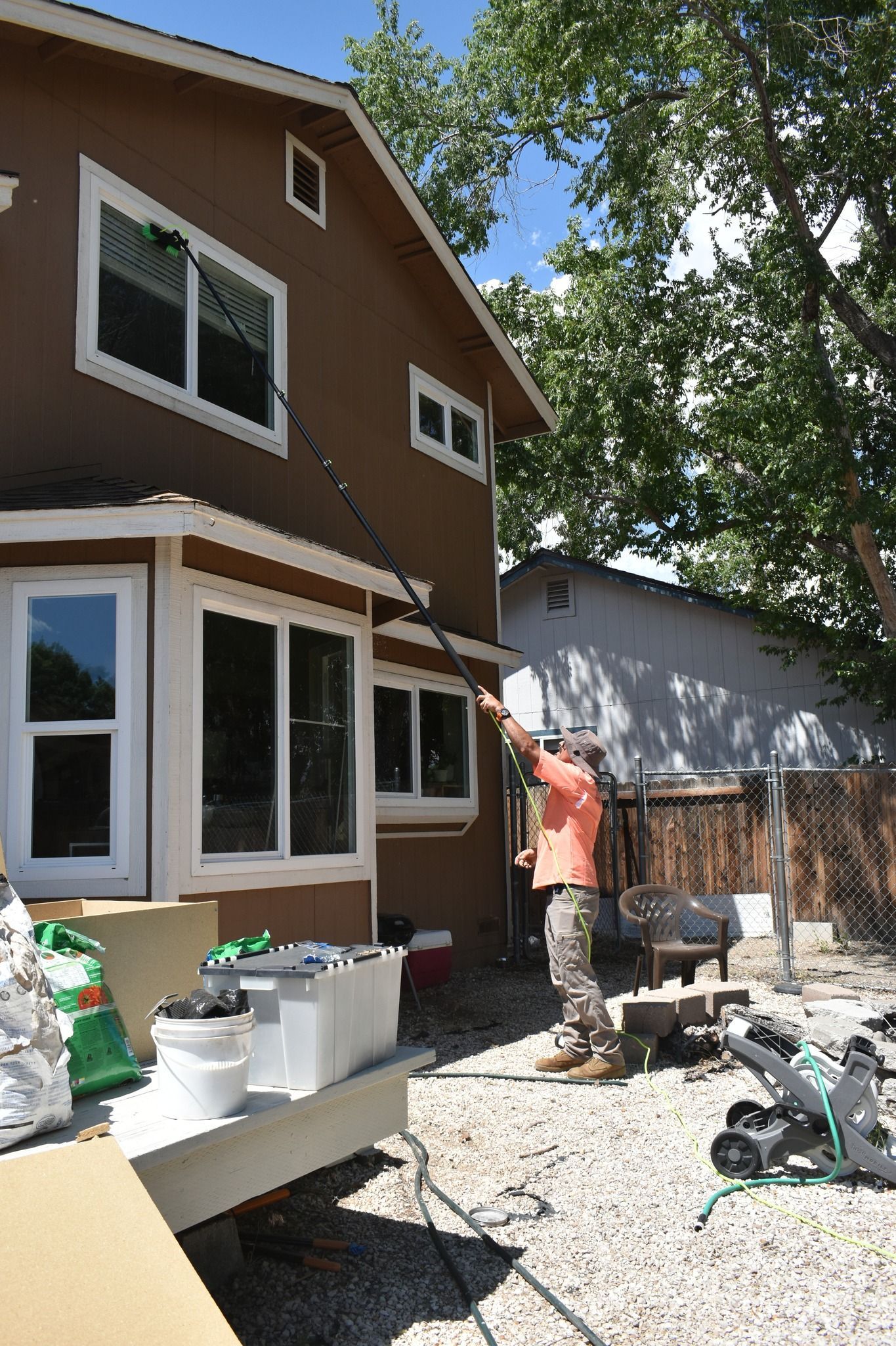 A person in an orange shirt uses an extended pole to clean the exterior second-story window of a brown house.