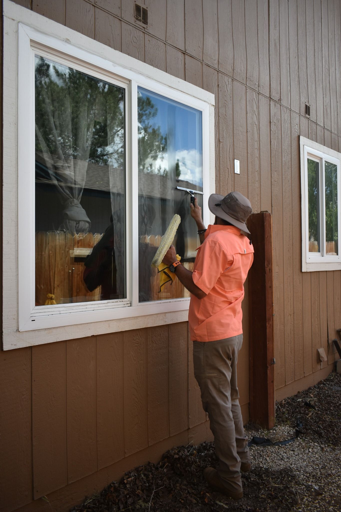 A person wearing a sun hat and peach shirt cleans an exterior residential window with a squeegee against brown siding.