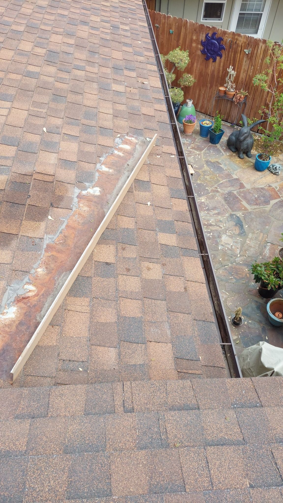 A view from a roof showing a long, wooden straight edge resting on brown shingles near a roof valley and a backyard patio.