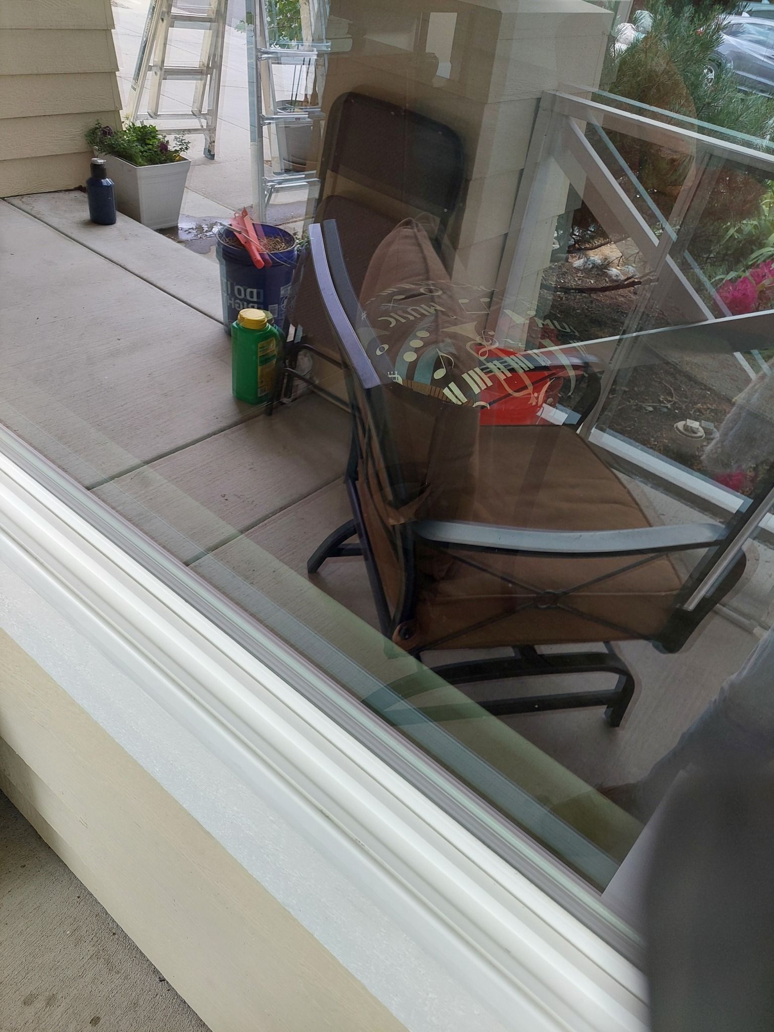 A porch view through a window, showing a brown patio chair, a potted plant, and containers on a concrete floor.