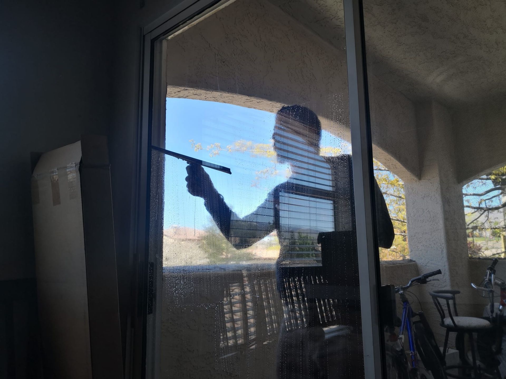 A person cleans a glass sliding door with a squeegee, with a balcony and outdoor scenery visible through the window.