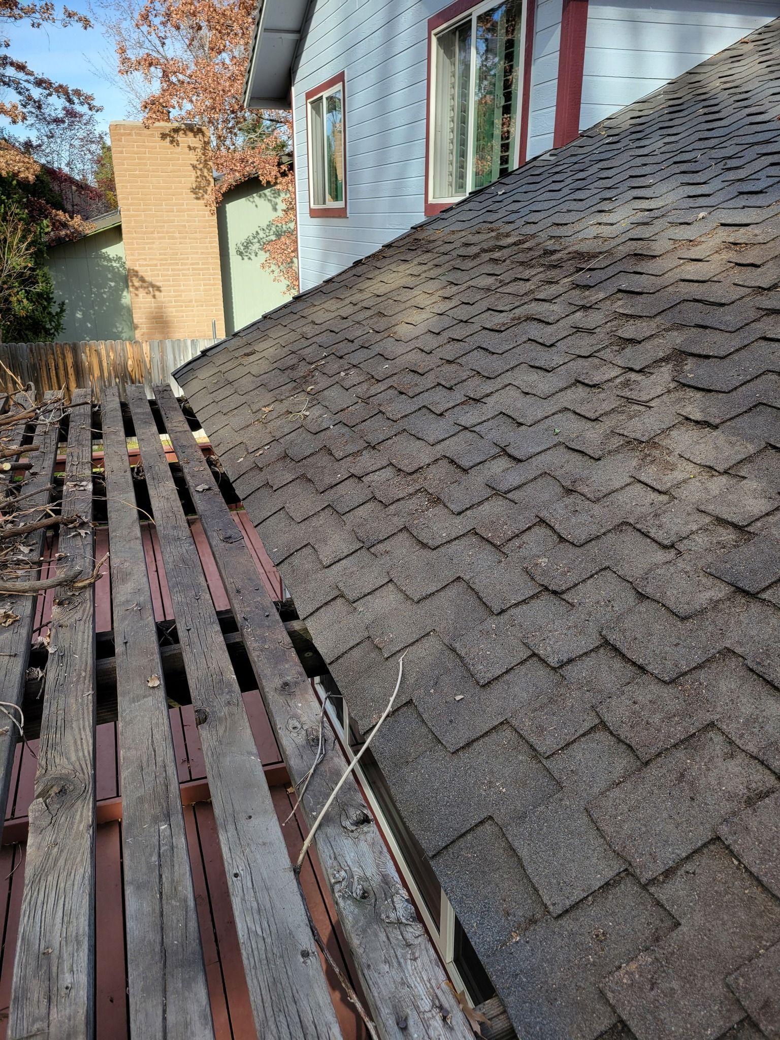 Roof section with dark shingles beside a stripped wooden frame on a sunny day.