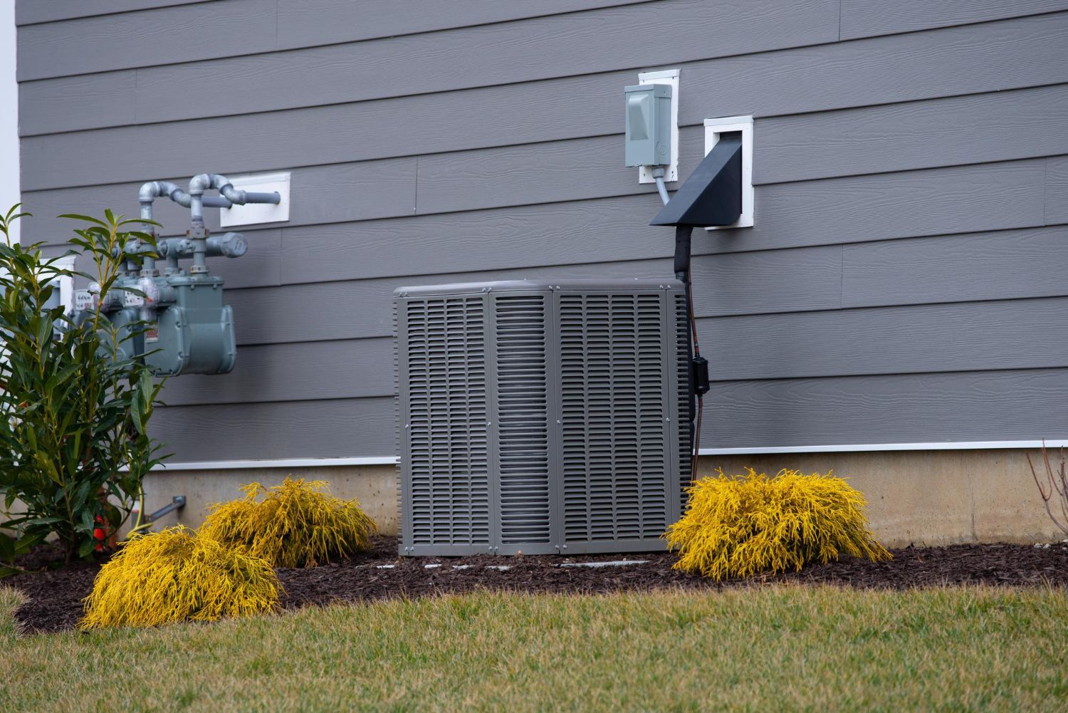 Outdoor utility meters and vent units on a gray house exterior with yellow shrubs