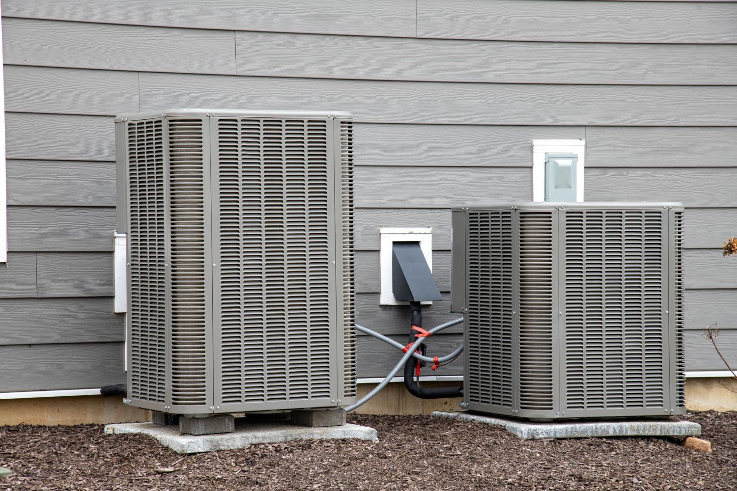 Two outdoor HVAC units beside a gray house wall on a gravel base.