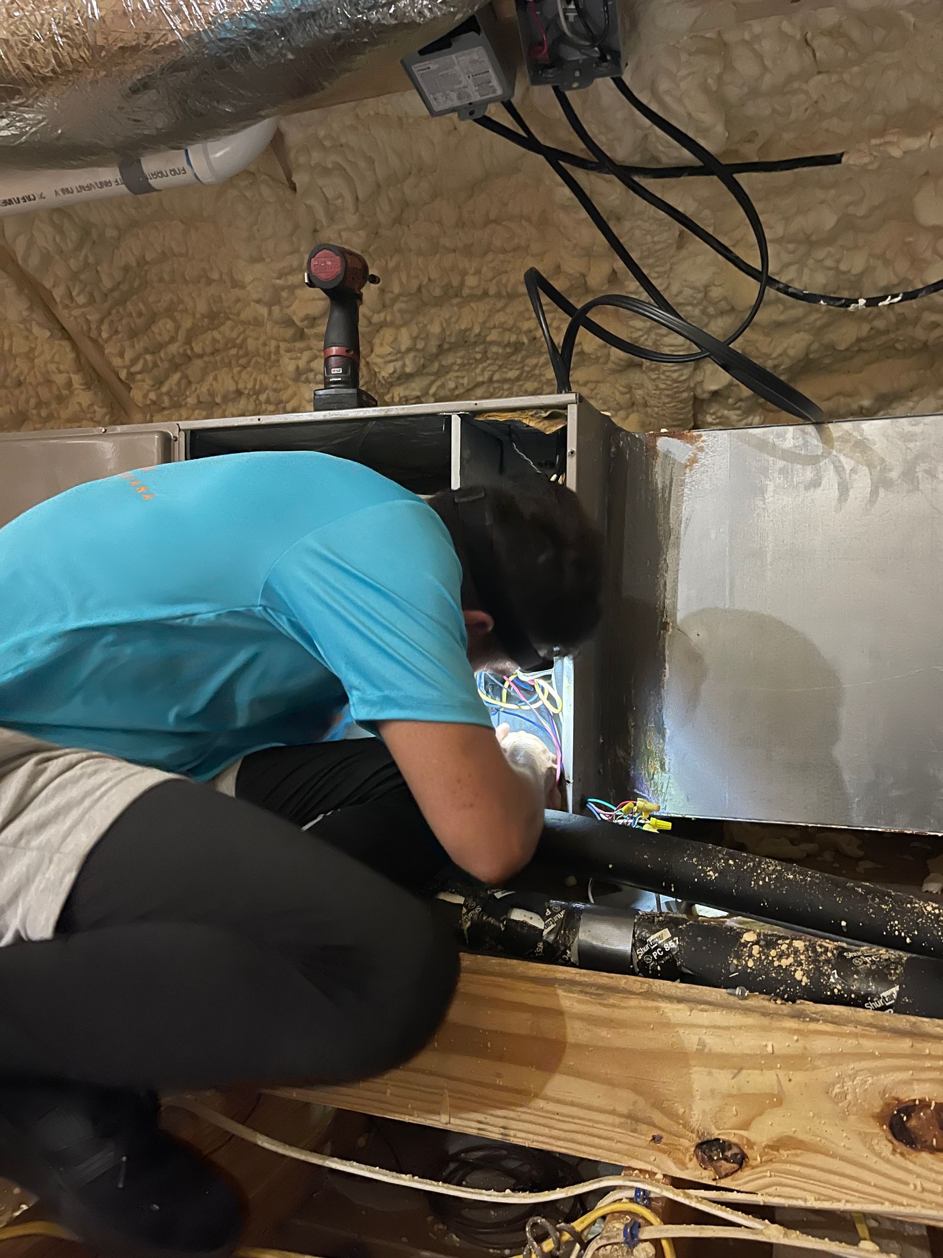 Person in blue shirt working inside a cramped crawlspace with pipes and wiring.