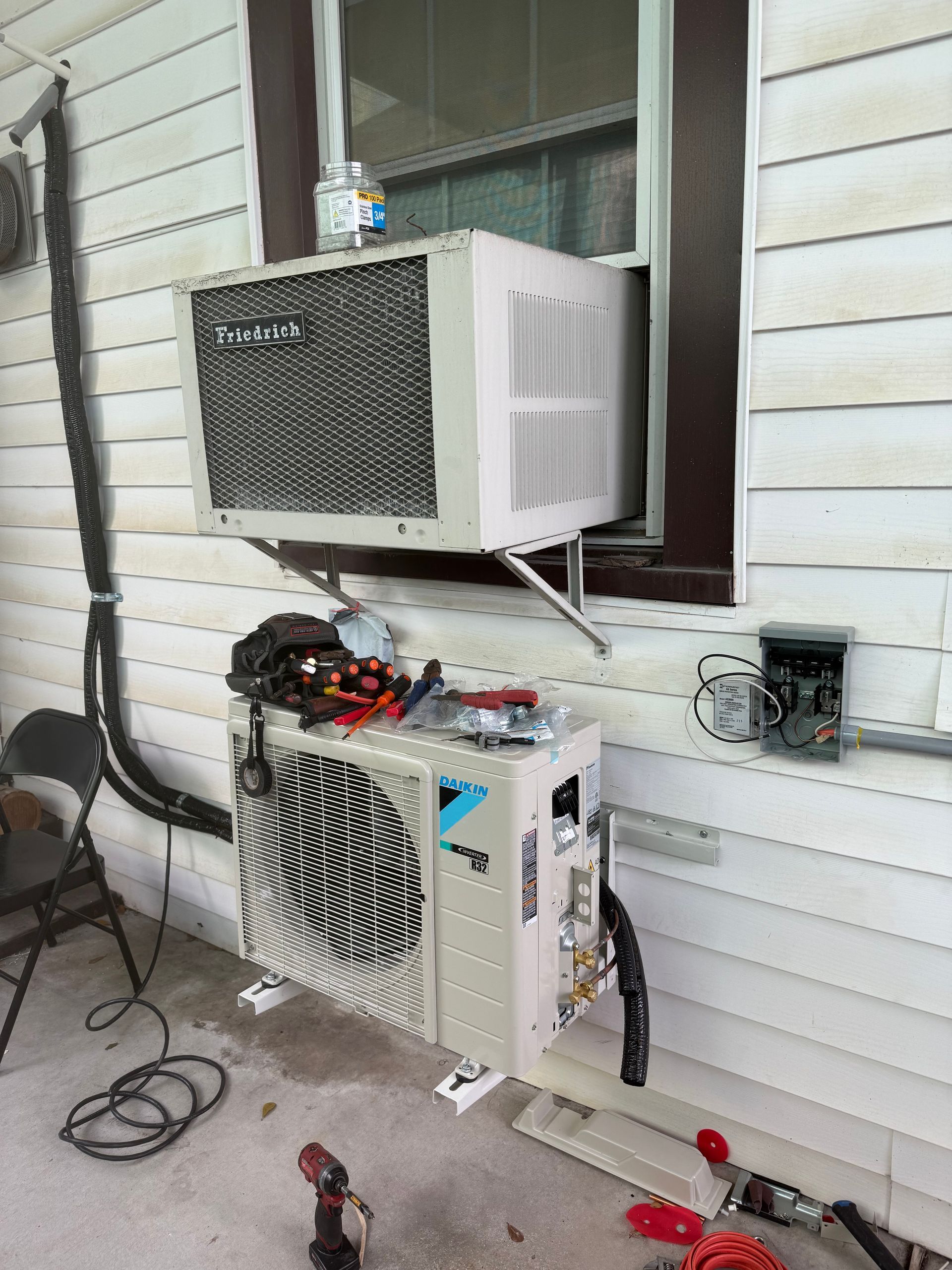 Two window AC units mounted outside a house, with wires and tools on the ground.