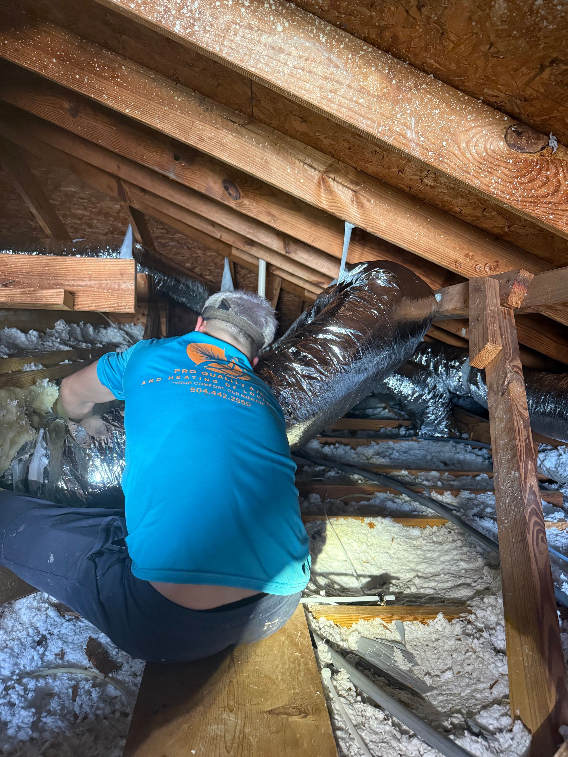 Person in blue shirt kneeling in an attic, working on insulation between exposed wooden rafters.