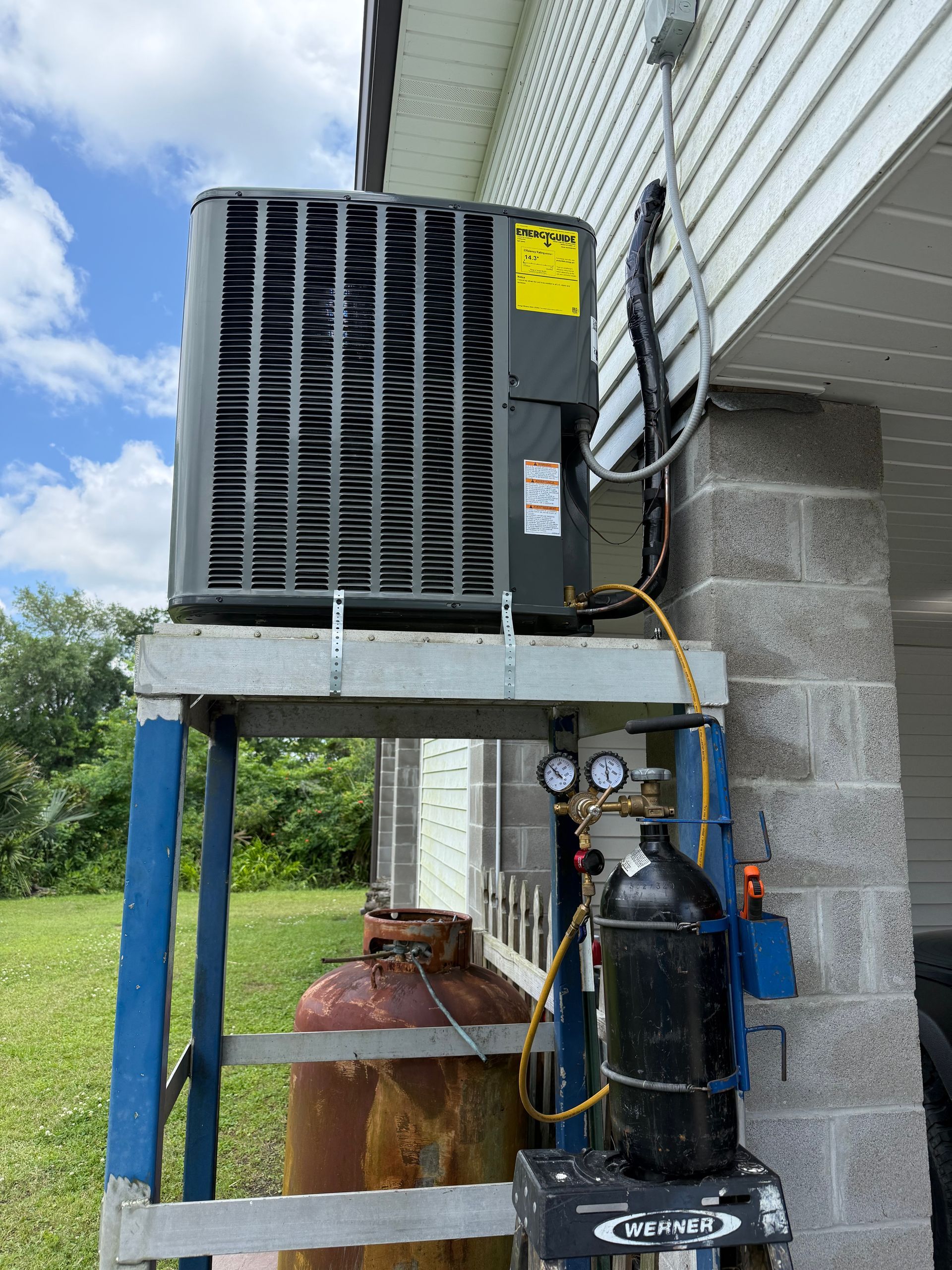 Outdoor HVAC unit on a raised platform beside a building, with gauges, tanks, and piping underneath.