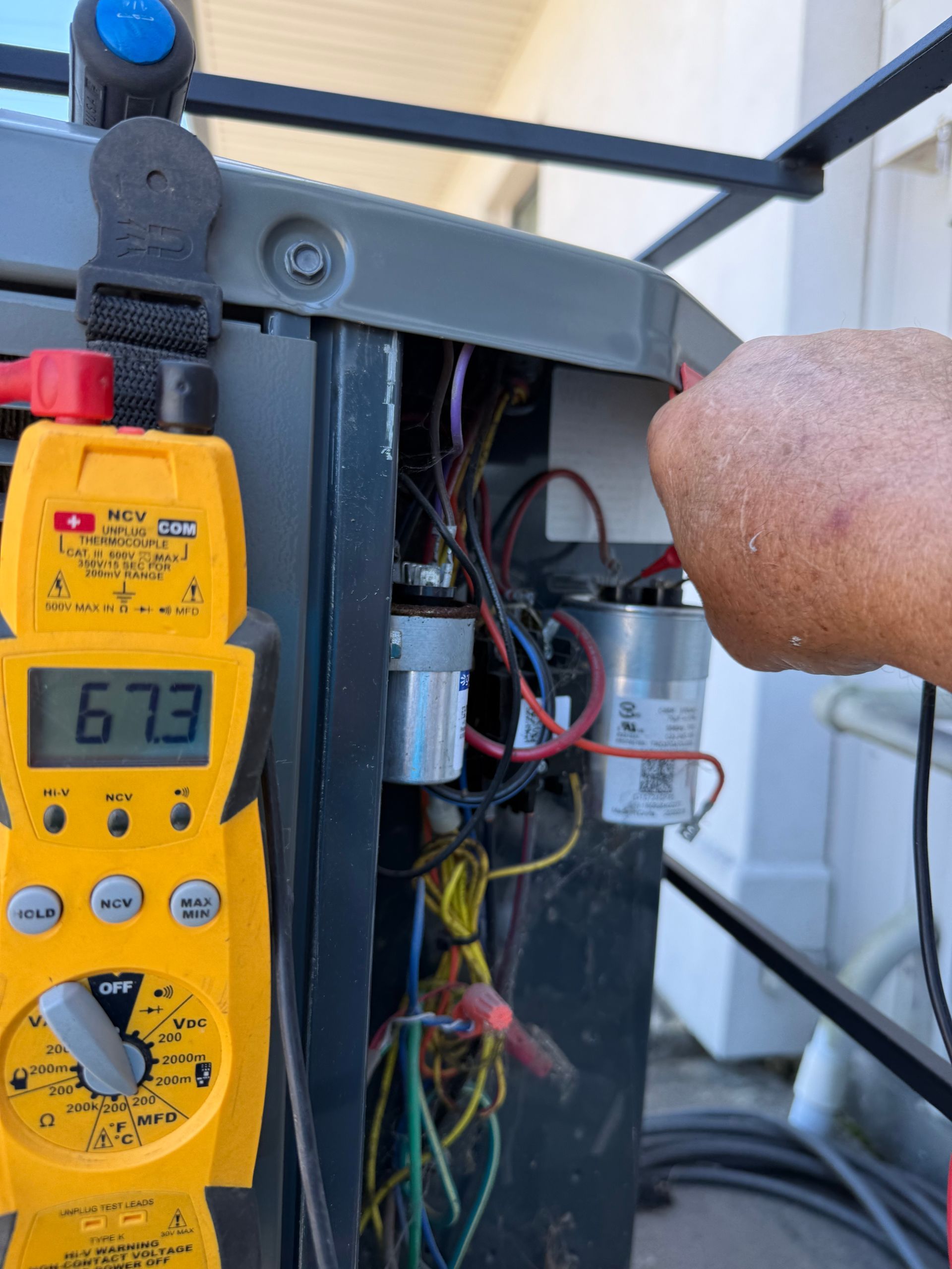 Yellow digital multimeter testing wiring inside an open electrical panel, with a hand reaching in.
