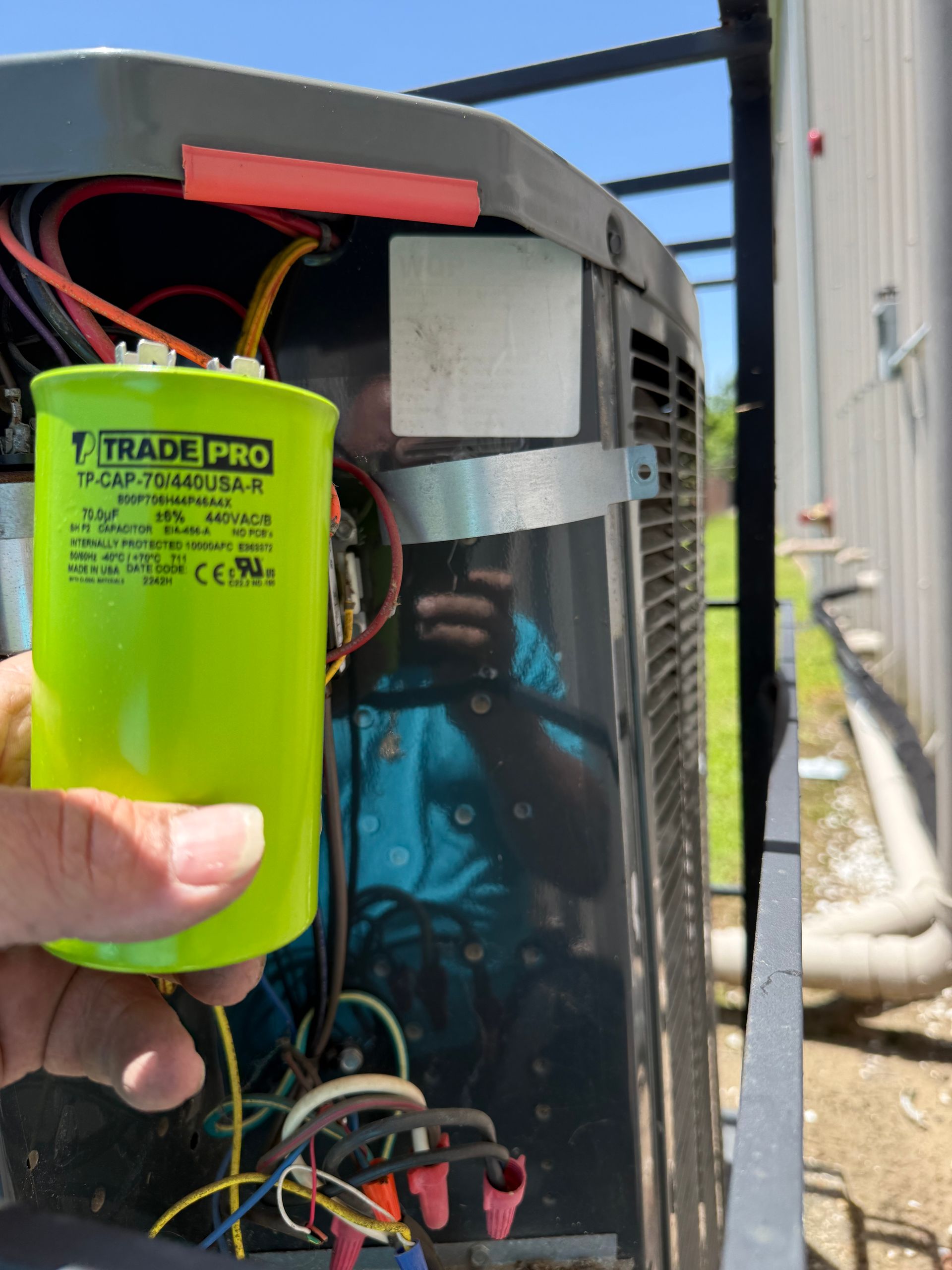 Hand holding a green Trade Pro capacitor inside an open HVAC unit with exposed wiring