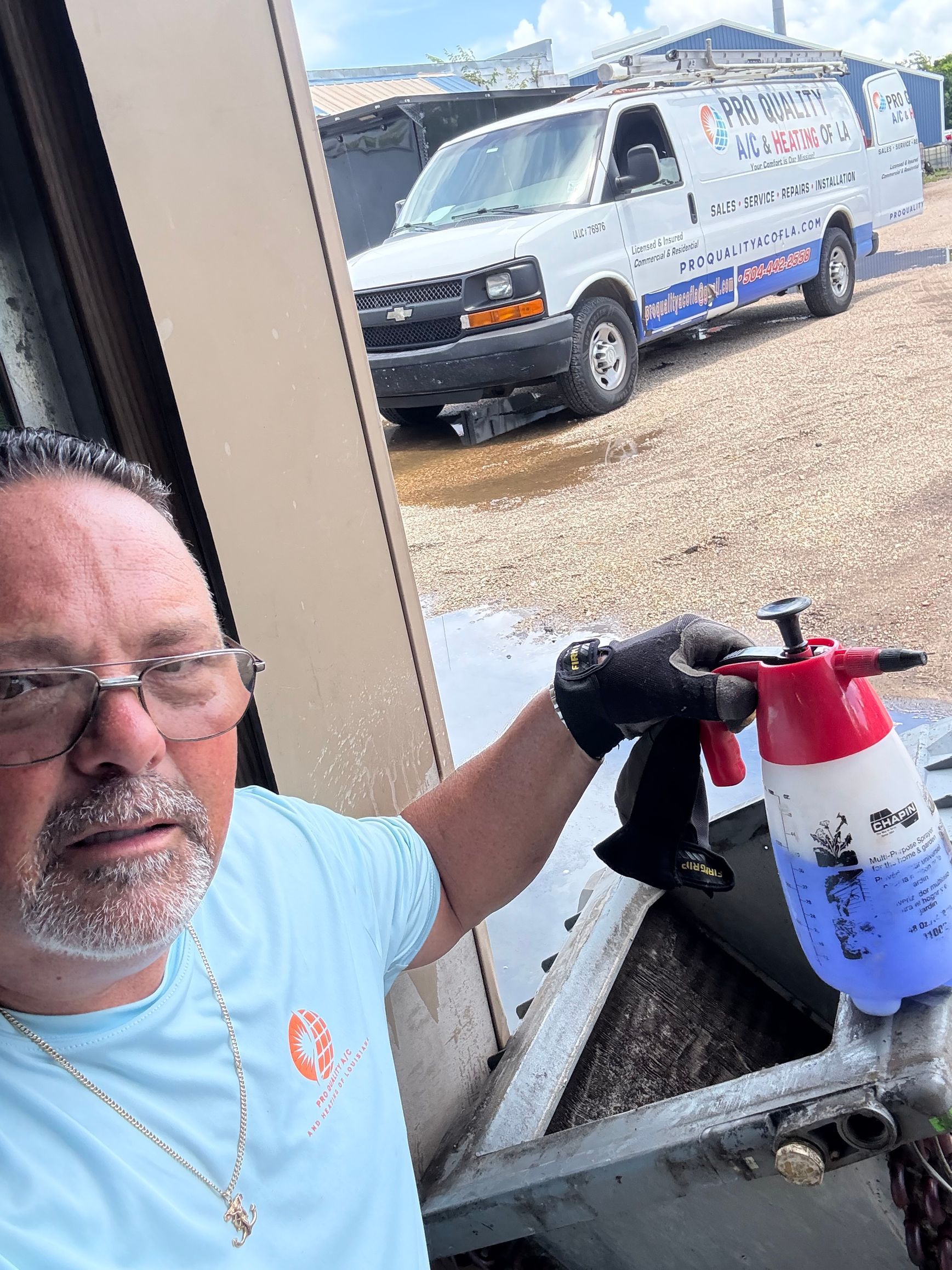 Man spraying equipment with a red-and-white sprayer near a workbench, with a utility van outside.