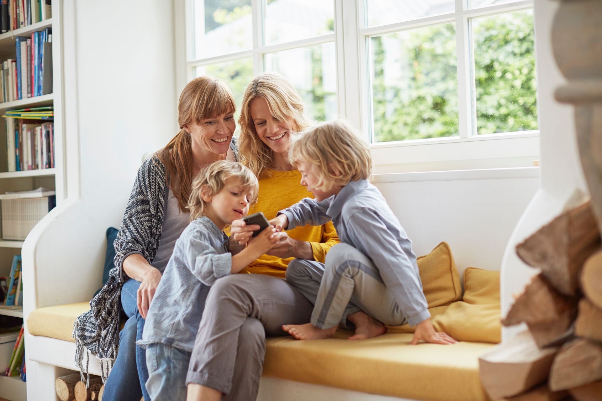 Four people sit on a yellow sofa, reading a book together by a bright window.