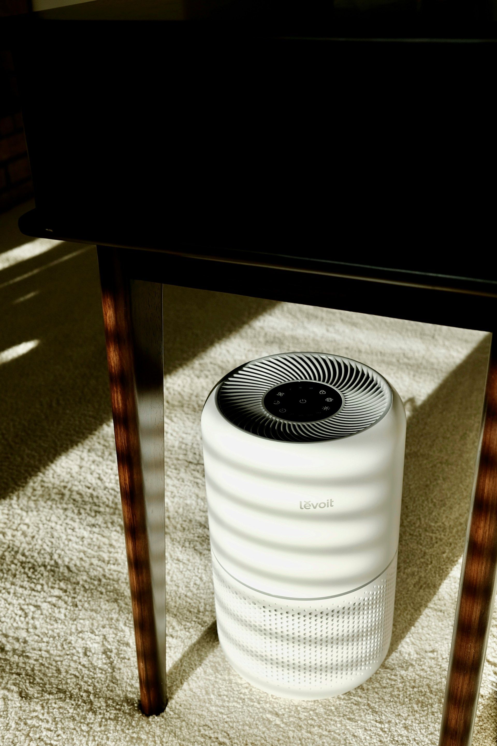 White cylindrical air purifier under a table, with a black vented top and textured lower section.