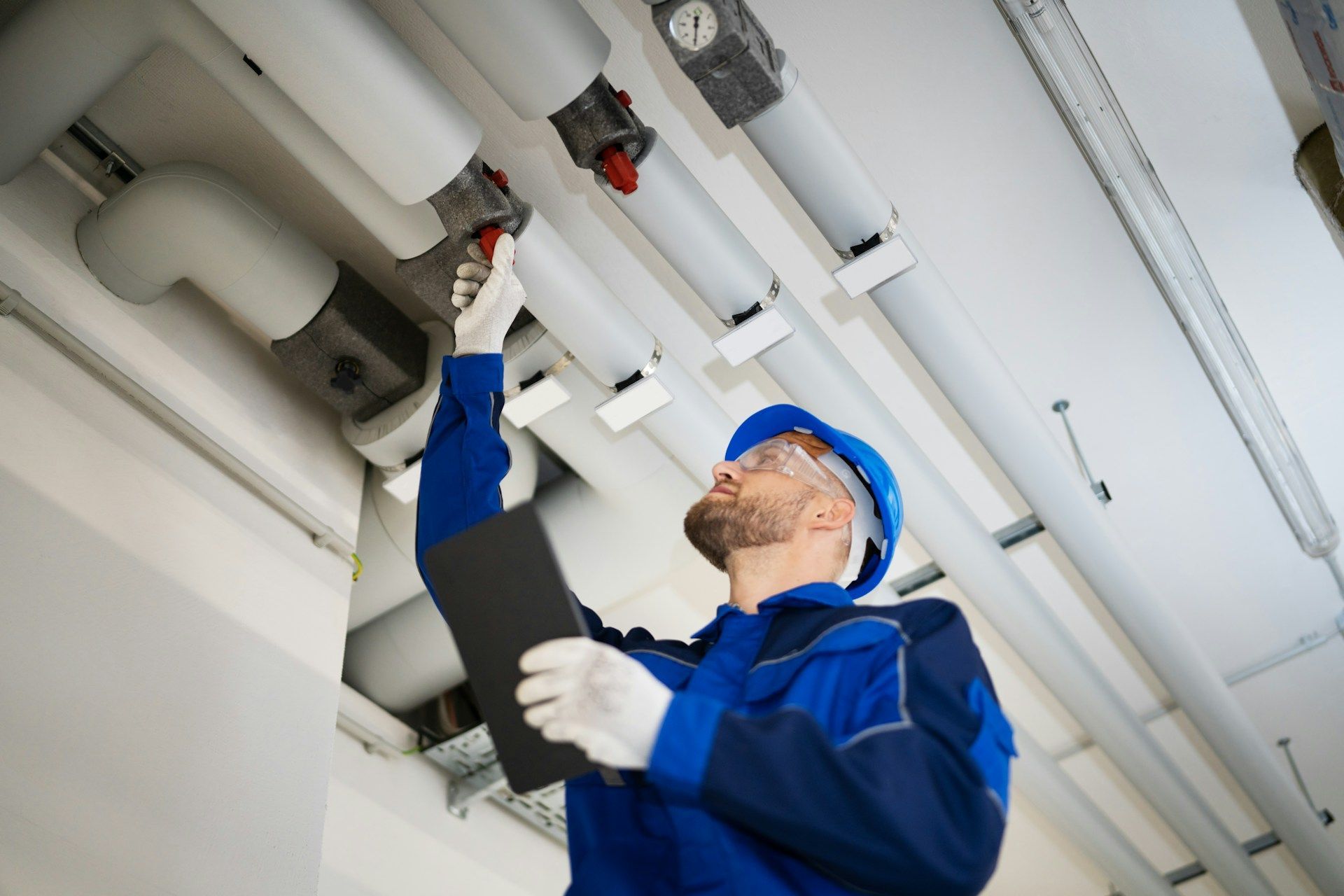 Worker in blue safety gear inspecting overhead pipes with a wrench in a utility room