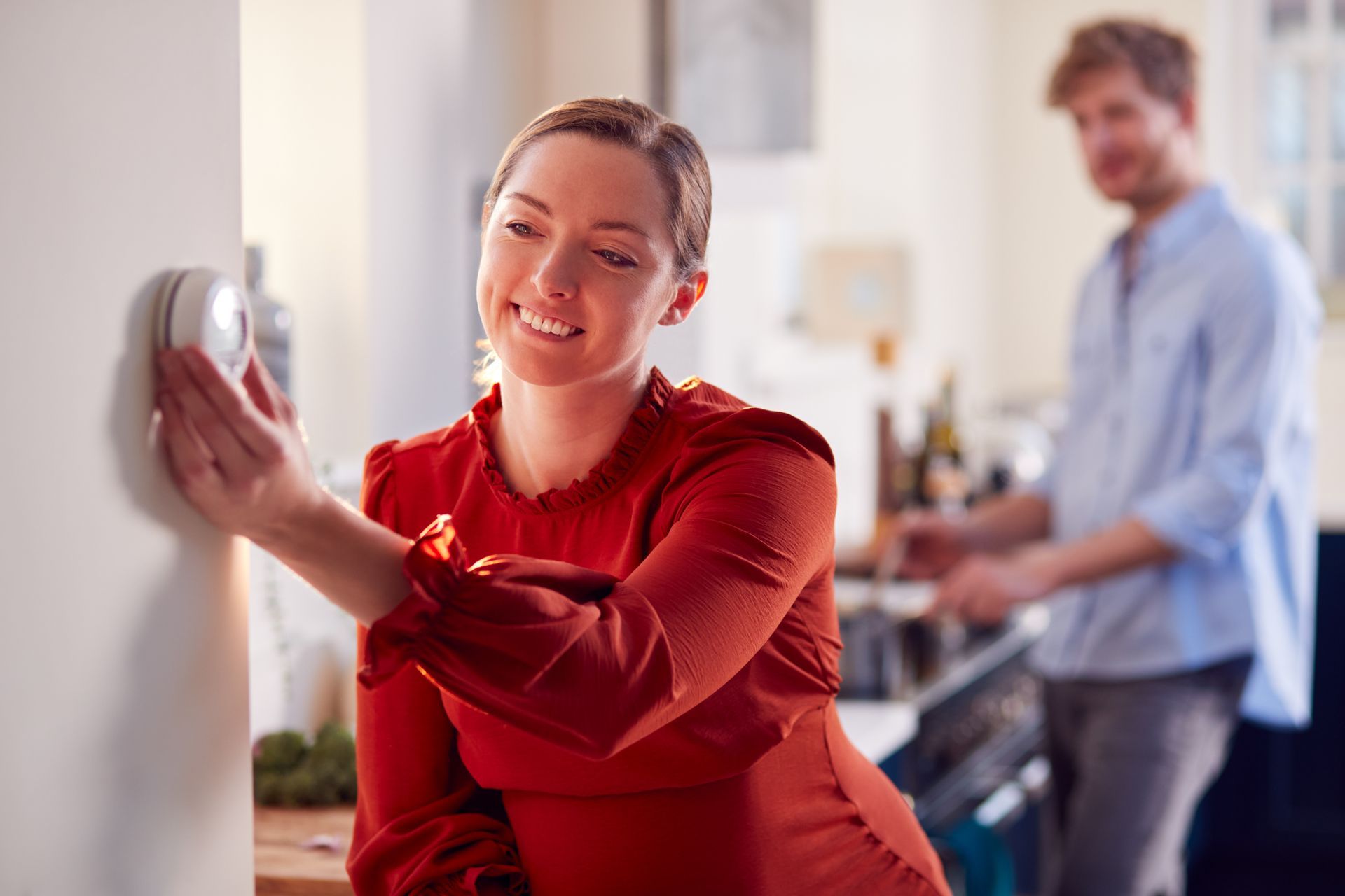Woman in a red shirt adjusting a wall thermostat, with a man standing blurred in the background.