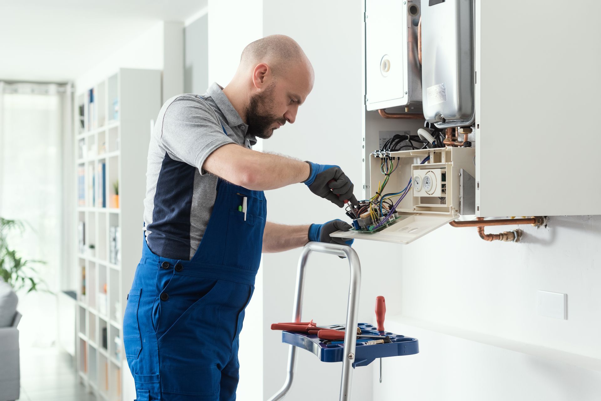 Technician in blue overalls repairs a wall-mounted boiler in a bright home kitchen
