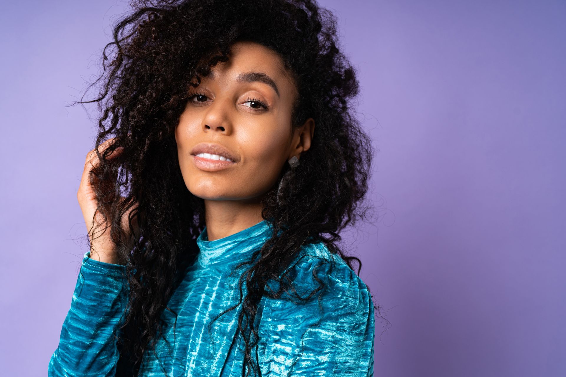 A woman with curly hair is holding her hair in front of a purple background.