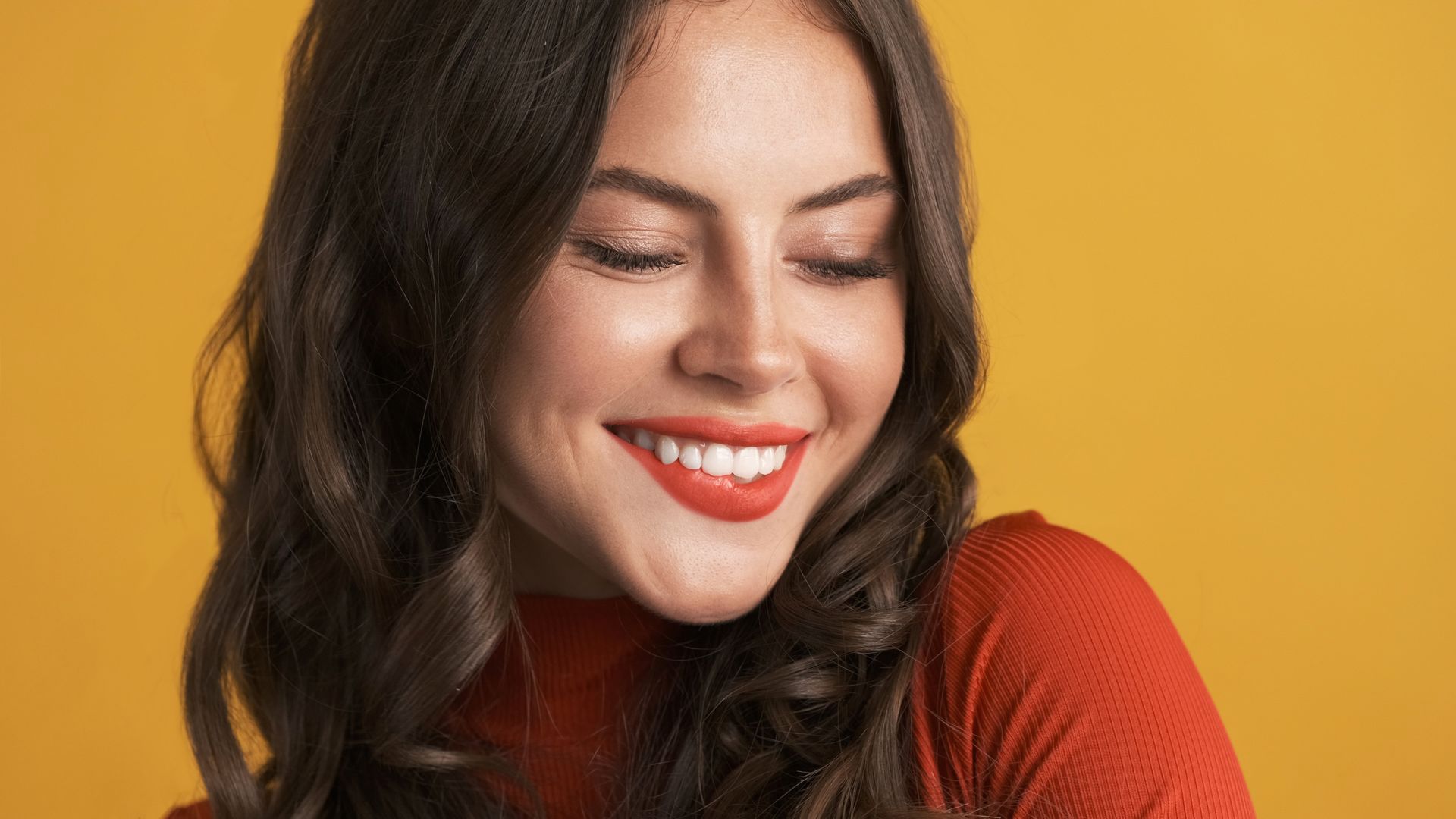 A woman is smiling with her eyes closed and wearing red lipstick.