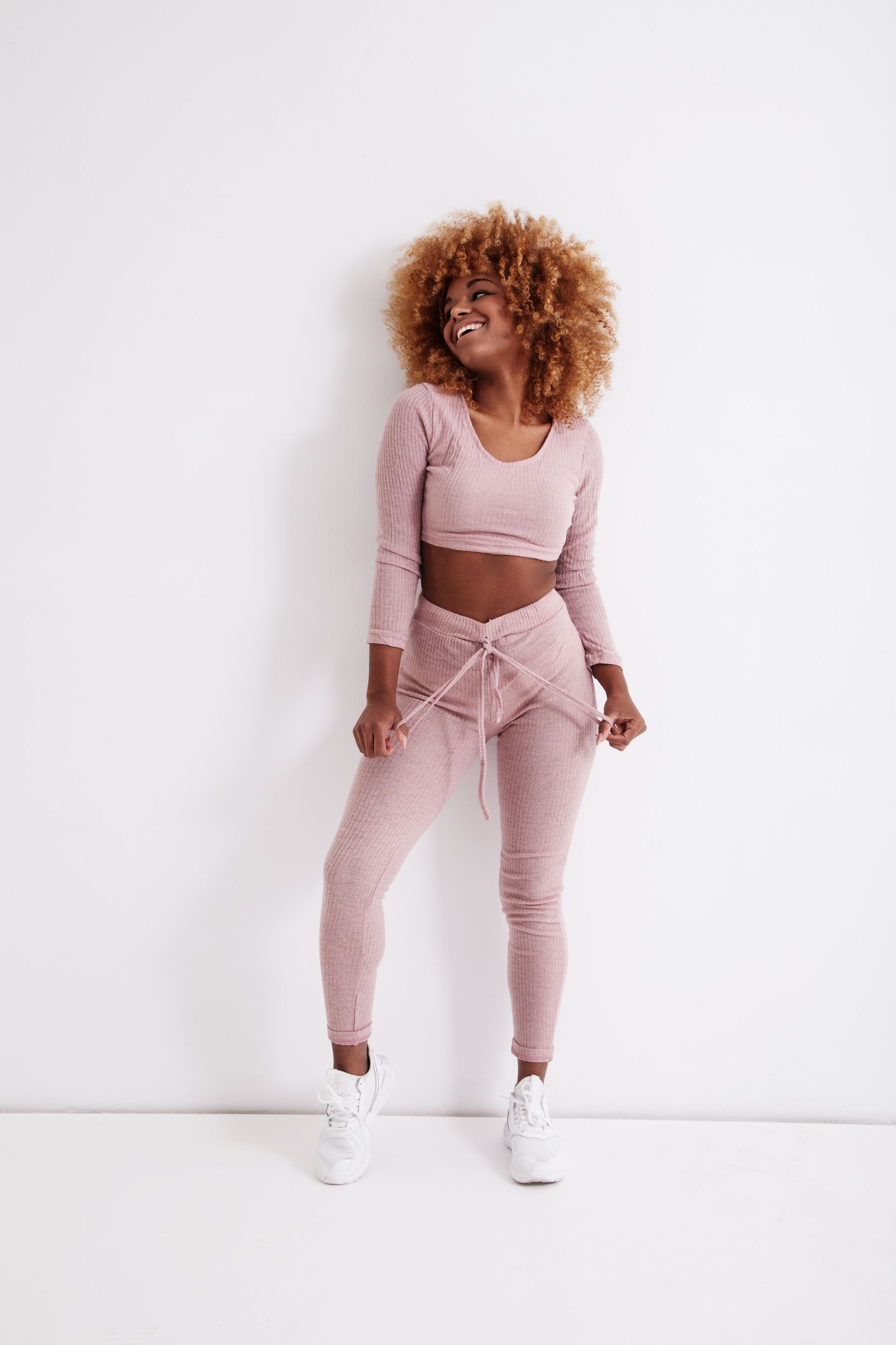 A woman in a pink crop top and pink sweatpants is standing in front of a white wall.