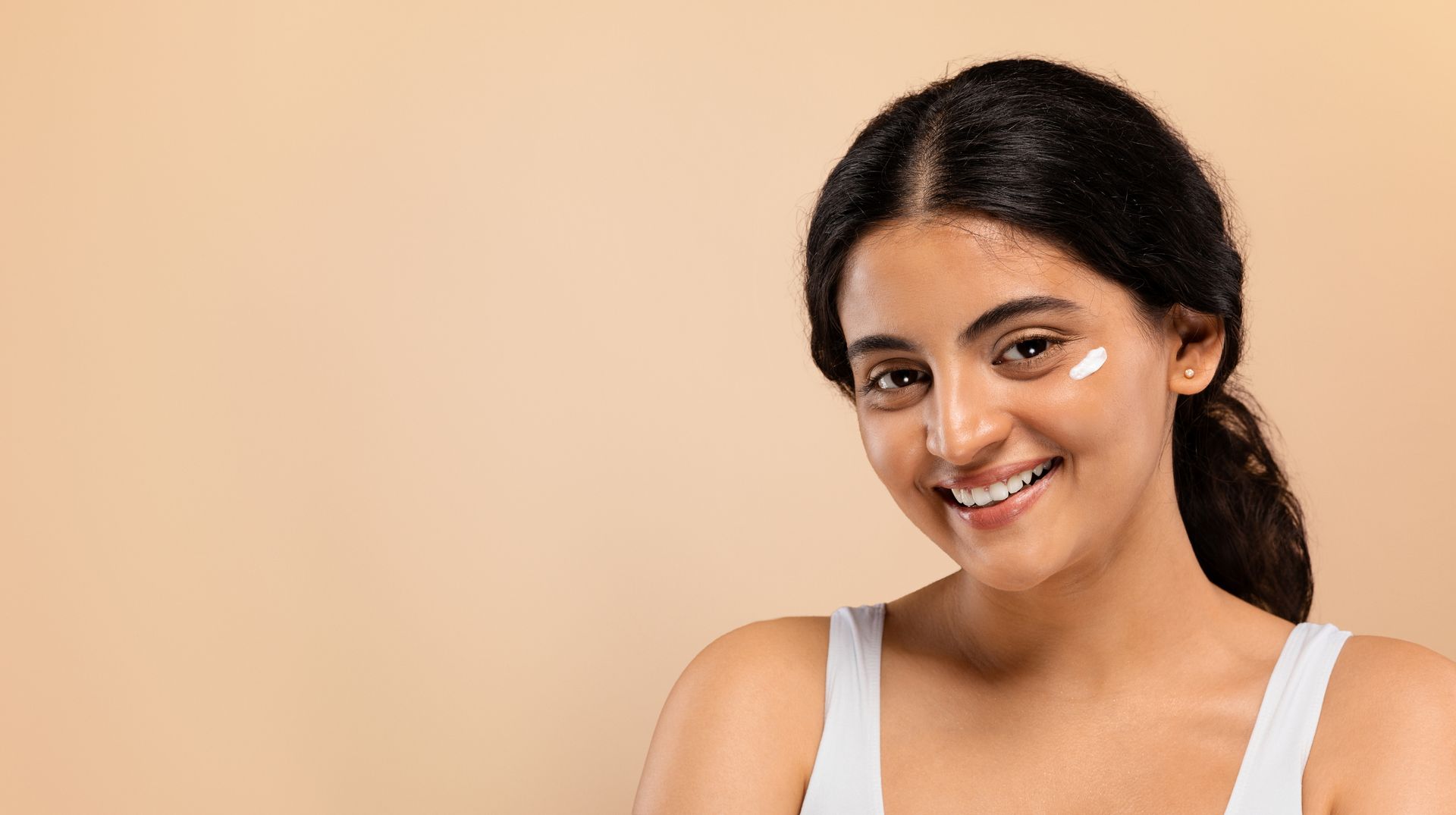 A woman is smiling and applying cream to her face.