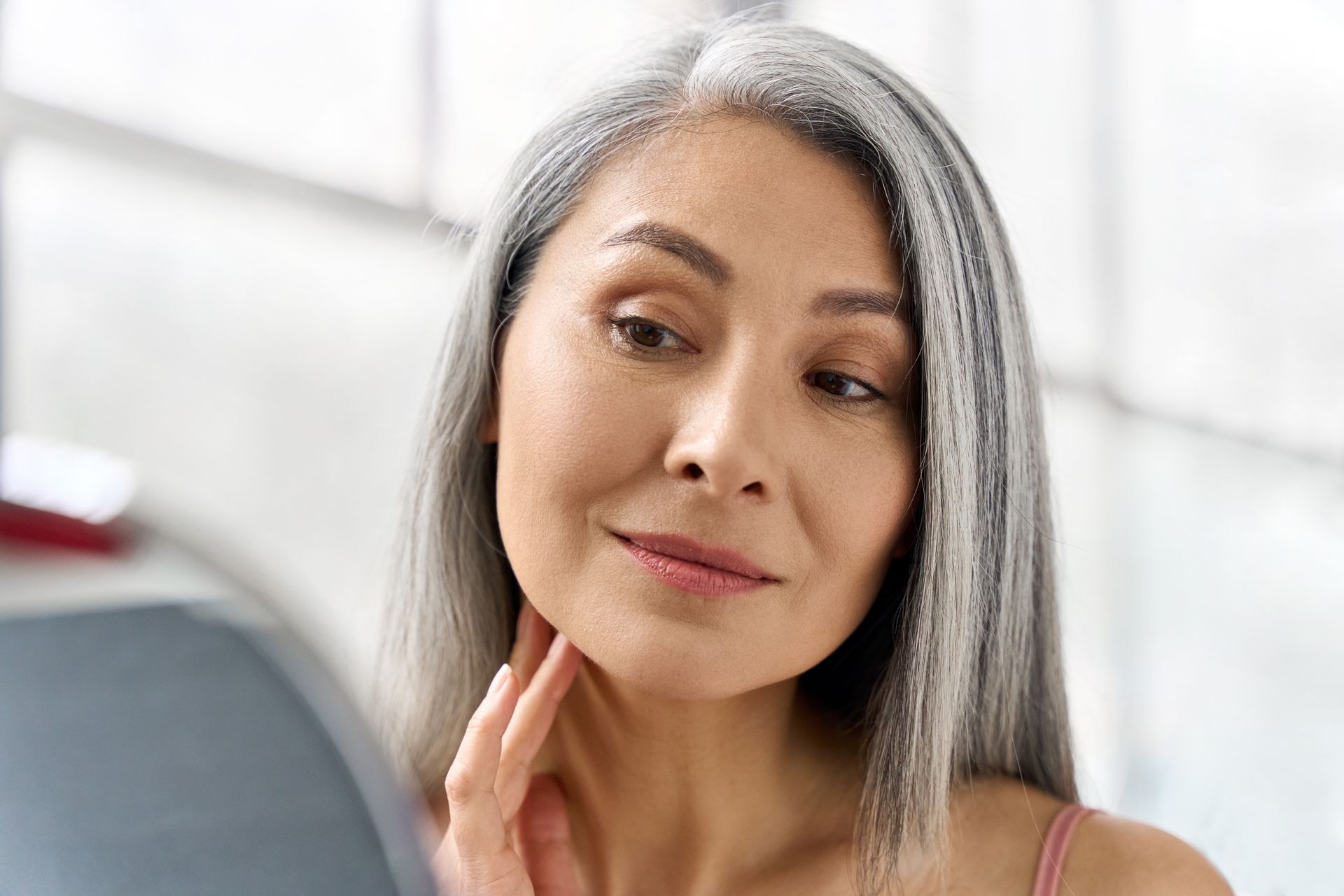 A woman with gray hair is looking at her face in a mirror.