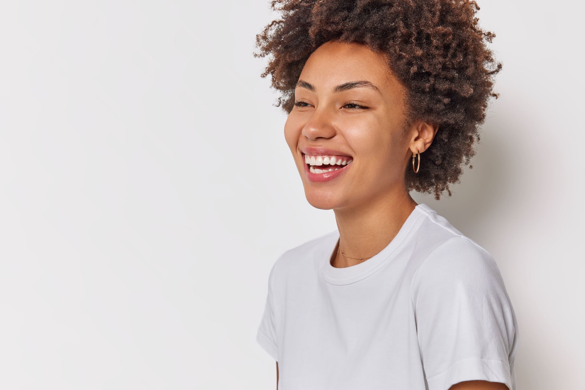 A woman with curly hair is smiling and wearing a white t-shirt.