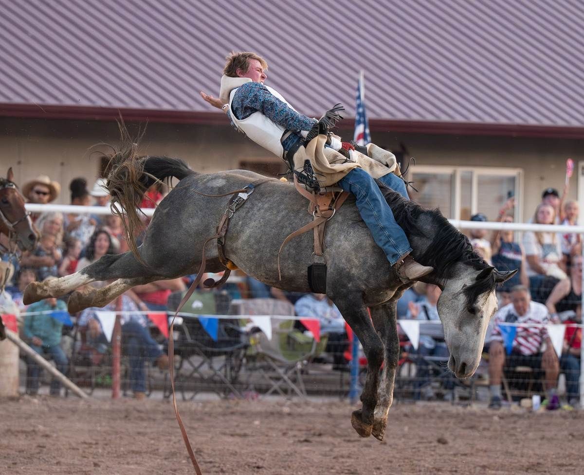 A rodeo rider on a bucking gray horse, airborne. Rider wears a vest and jeans, in an outdoor arena.