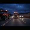 Two red trucks and cars driving on a road at dusk. Dark blue sky overhead.