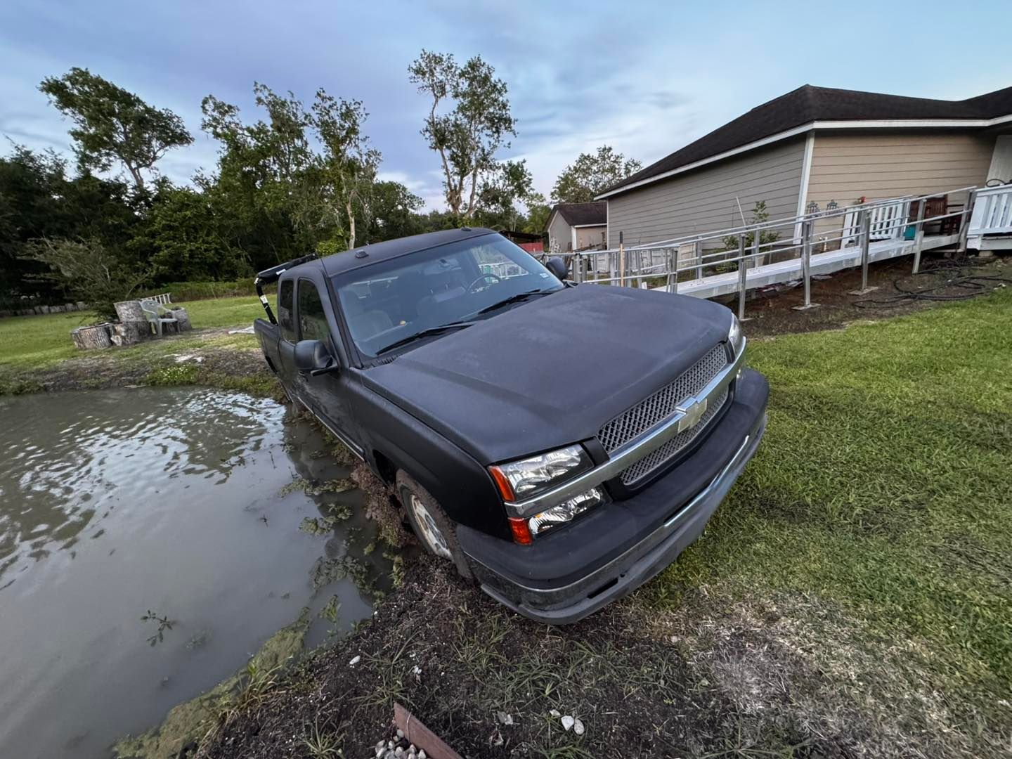 Black truck stuck in muddy water near a house with a ramp; overcast sky.