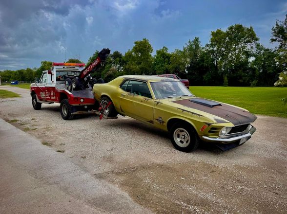 Tow truck towing a classic yellow and brown Mustang on a gravel road. Overcast sky, trees in the background.