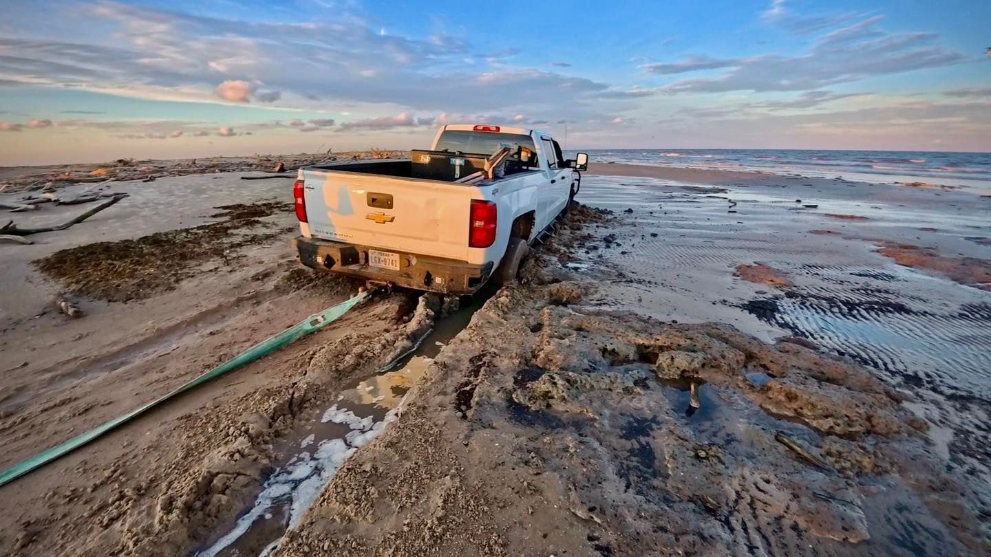 White pickup truck stuck in muddy sand near a body of water, with a hose trailing away.